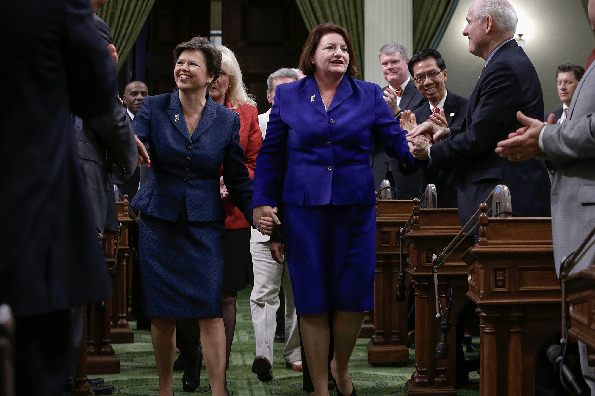 Two people walk hand-in-hand down the center aisle of a legislative chamber as others around them stand and applaud. Both wear dark blue suits and smile warmly. The setting features ornate wooden desks and green carpeting, indicating a formal government space.