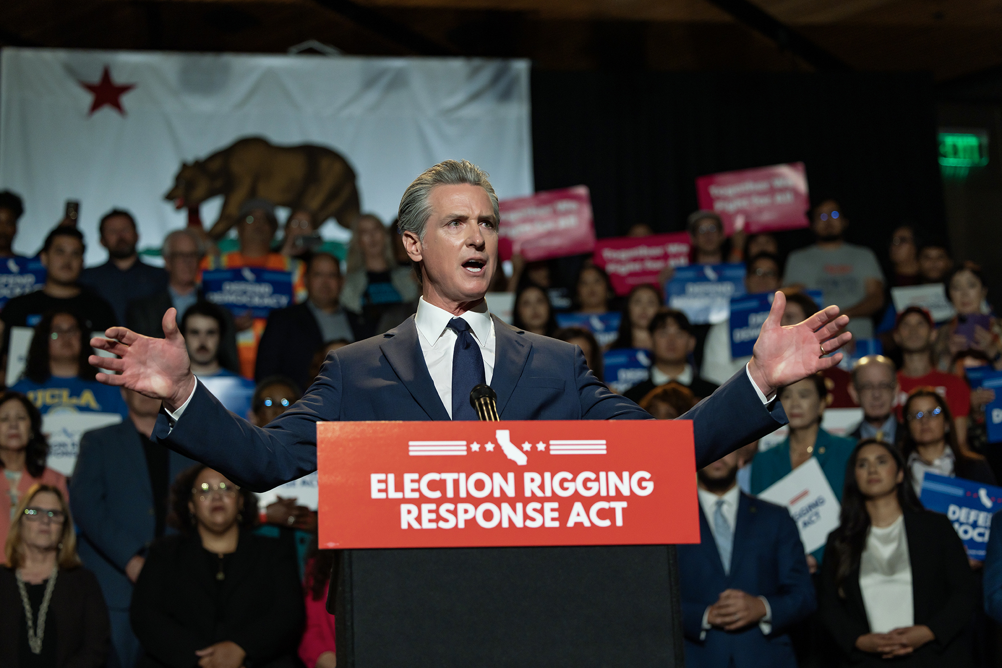 This image captures a close-up moment of the rally. The speaker at the podium is gesturing emphatically with arms outstretched, underscoring the intensity of the speech. The podium sign reads “Election Rigging Response Act,” and the California state flag is prominently displayed behind, reinforcing the political context. Supporters in the background hold signs with slogans such as “Defend Democracy.” The photo conveys a sense of urgency, leadership, and collective mobilization around protecting election integrity.
