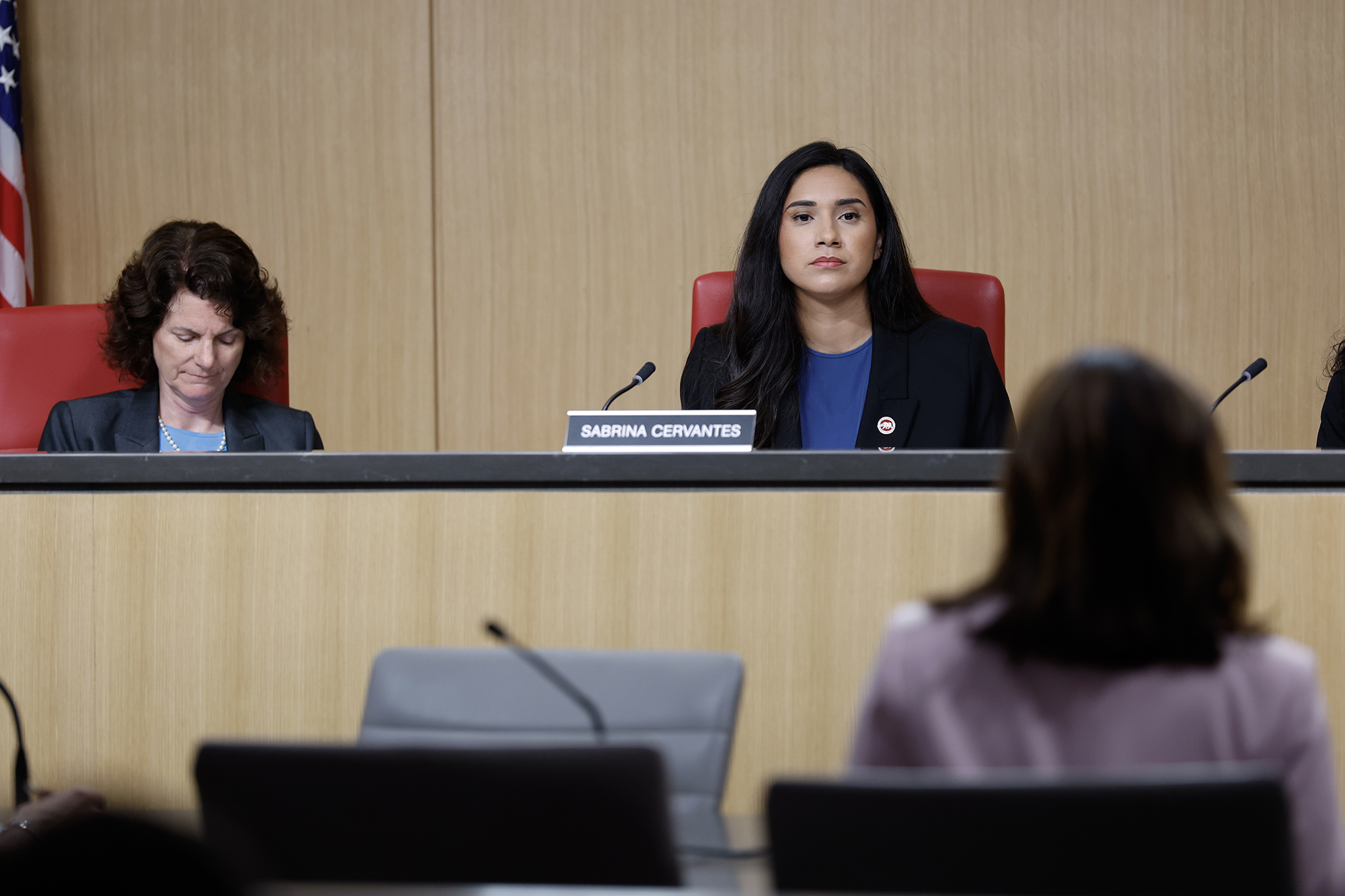Two people sit behind a panel desk in a hearing room. One looks down at notes while the other, seated in the center, looks forward with a neutral expression. A microphone and nameplate reading “SABRINA CERVANTES” are visible on the desk, with a blurred figure in the foreground facing them.