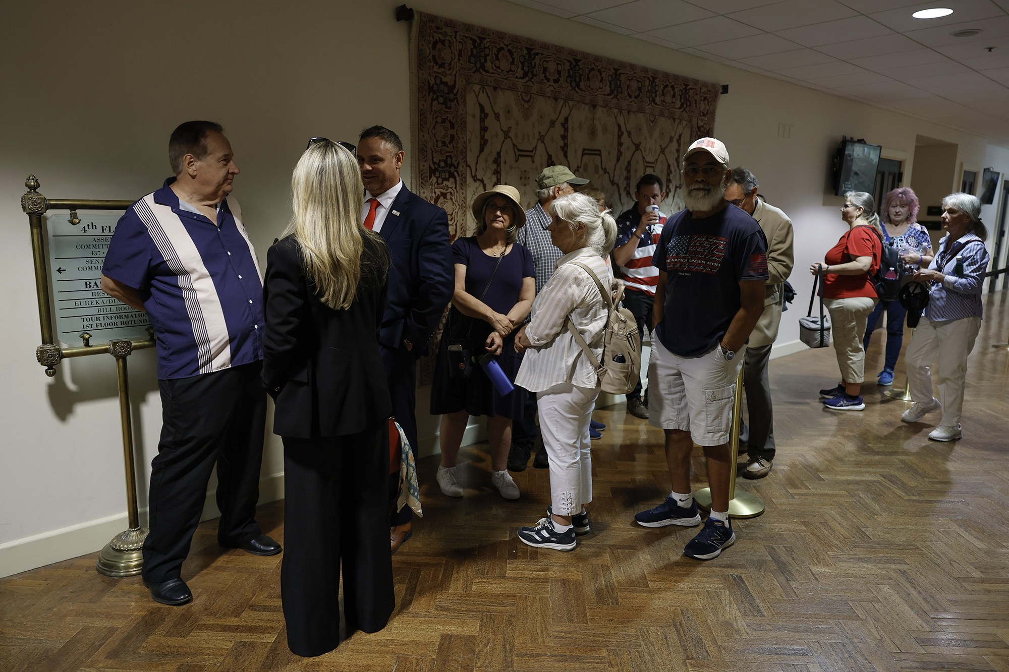 A group of people waits in line inside a government building hallway, some talking while others stand quietly.