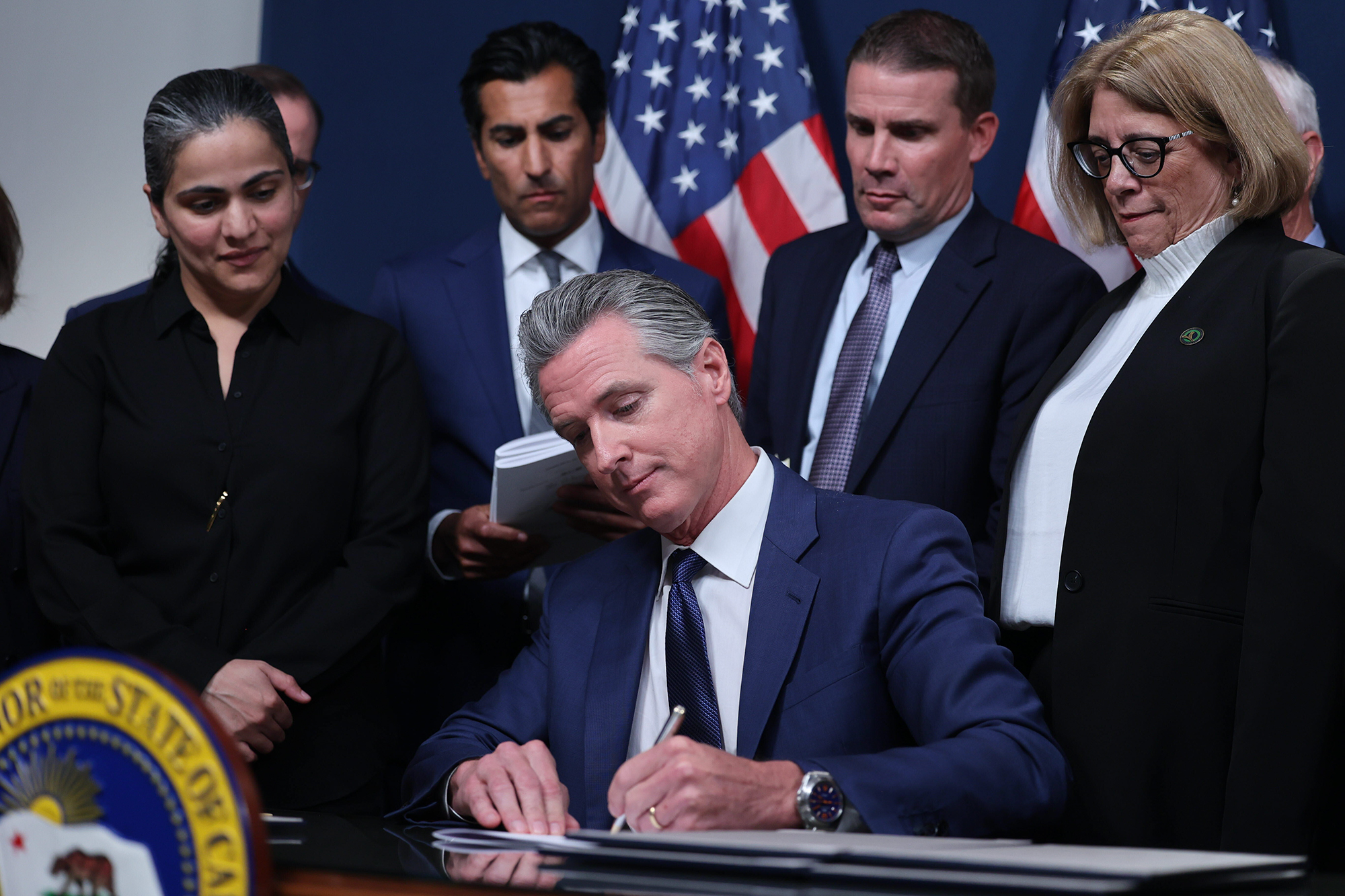 A lawmaker writes on a sheet of paper as they sit in front of a desk while surrounded by other lawmakers during a press conference.
