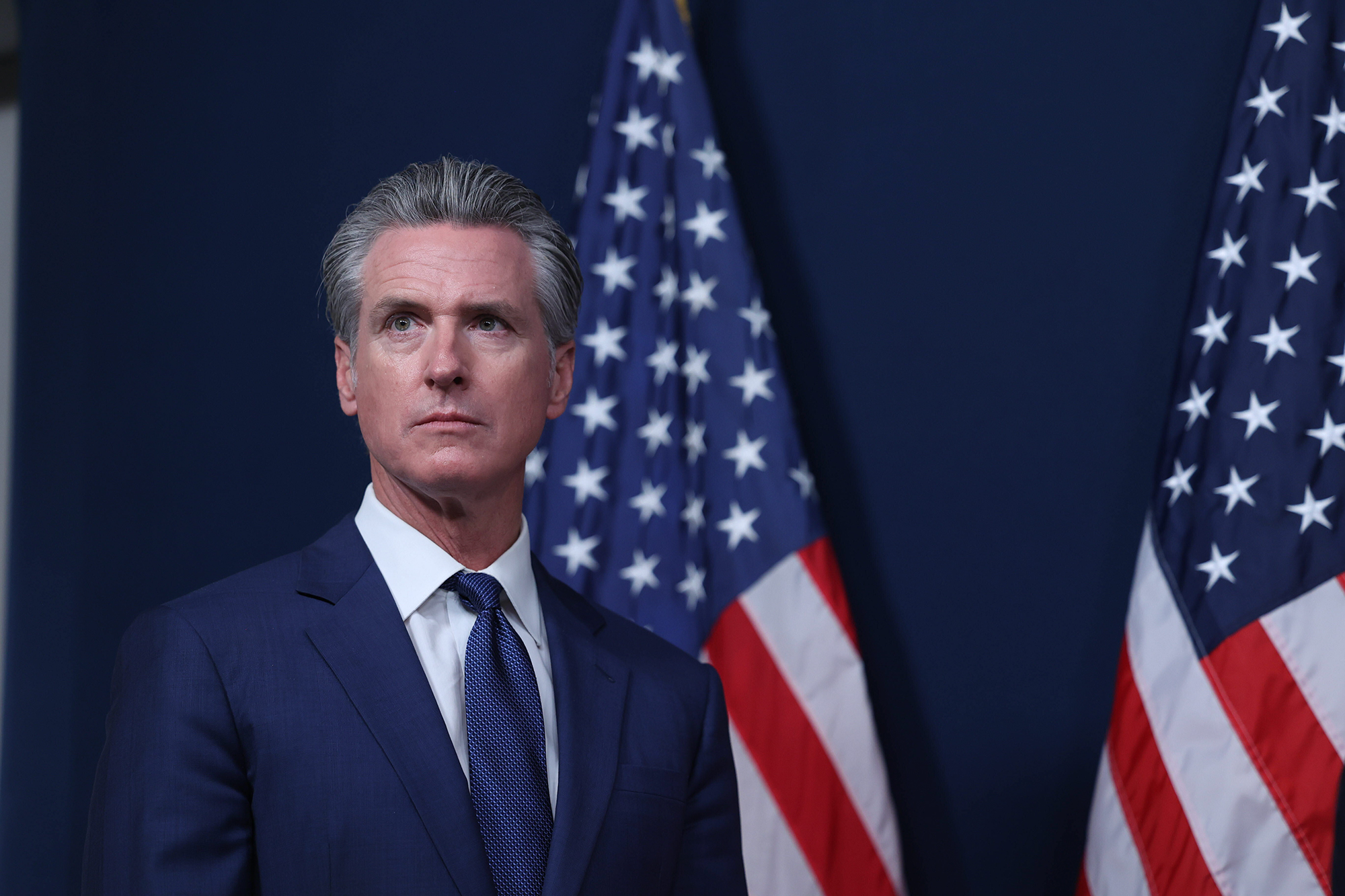 A person, wearing a blue suit, stands in front of two American flags while as they look straight off into the distance during a press conference.