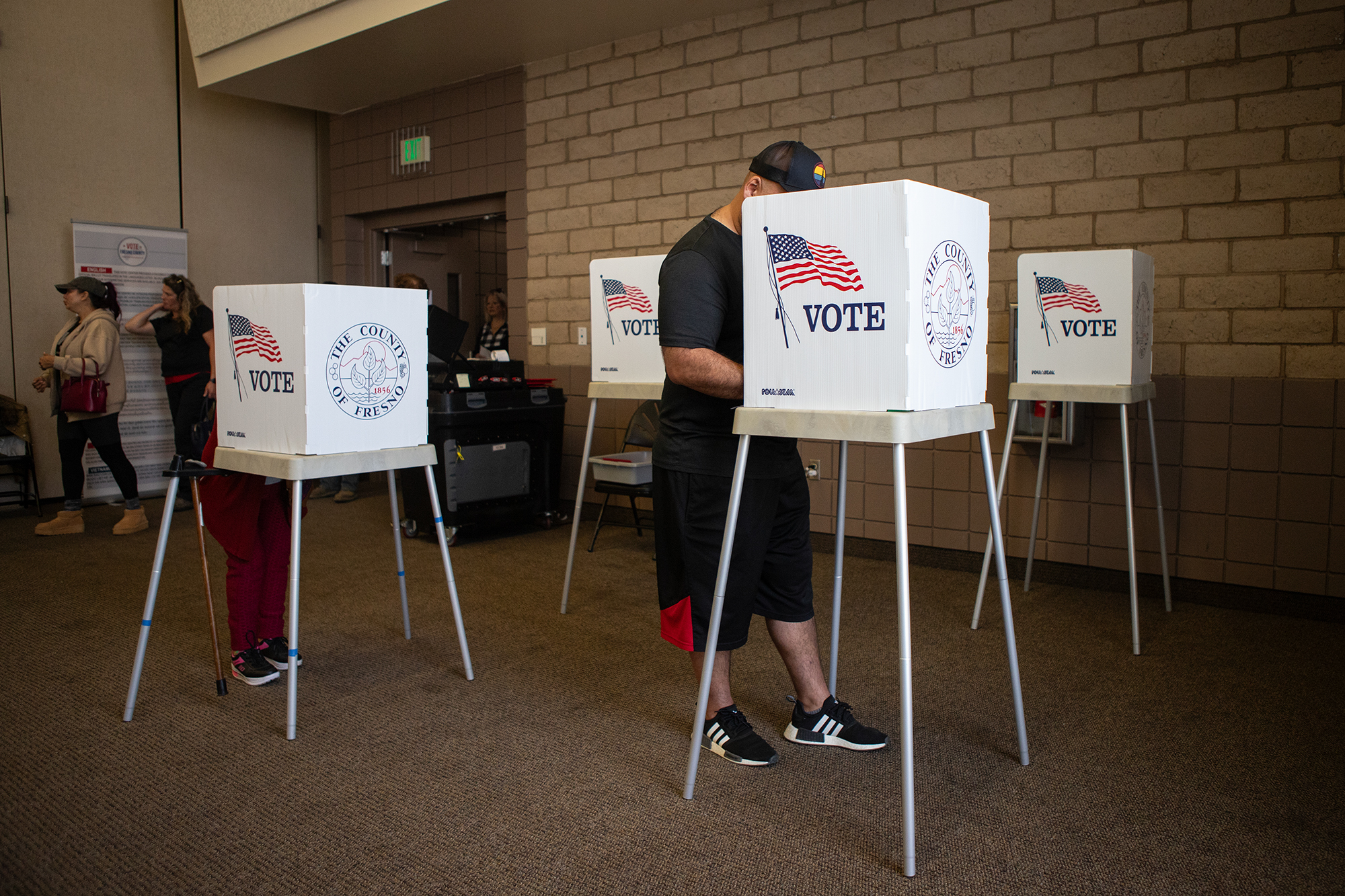 Four tall tables with white dividers on them that say "vote" next to an image of the American flag as people fill out their voting ballots. One person, in the center, wears a black hat, shirt and shorts, as they look down at their ballot.