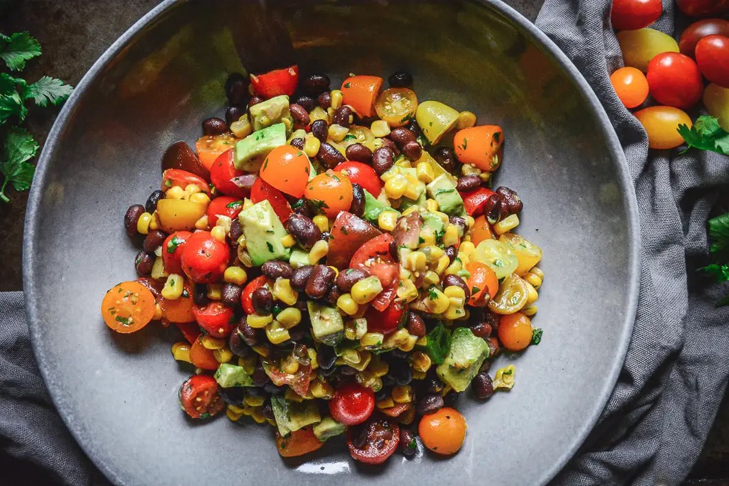 A delicious, fresh, and filling Mexican Tomato Salad with Black Beans, Corn, and Avocado in large bowl with grey napkin next to it