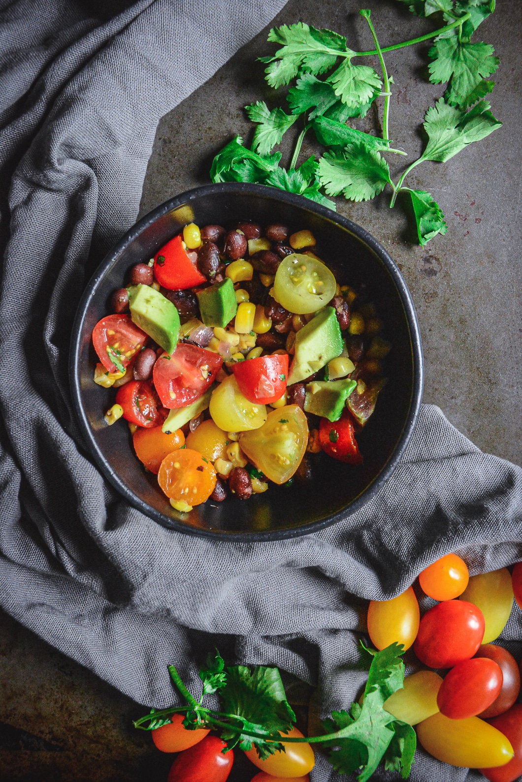 A delicious, fresh, and filling Mexican Tomato Salad with Black Beans, Corn, and Avocado in small black bowl with grey napkin