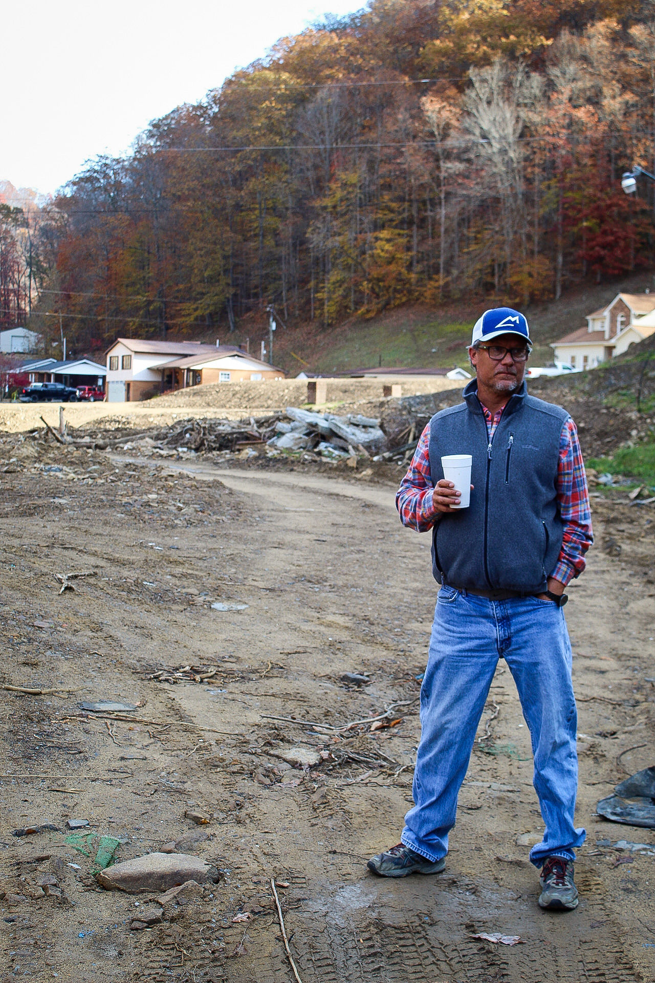 A man, Bart Chambers, stands on a muddy lot near houses that have been flooded out in Buchanan County