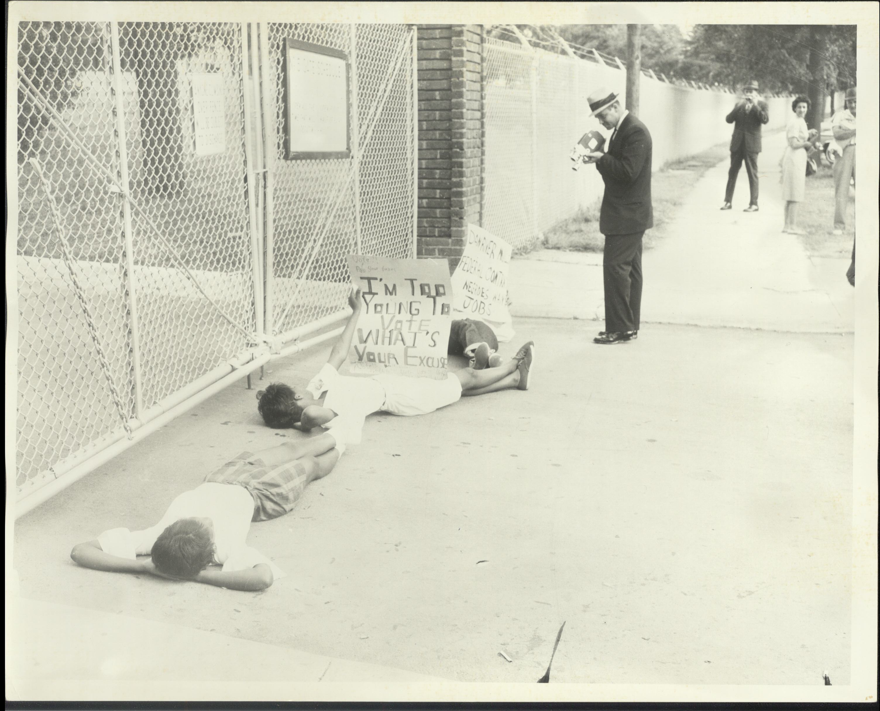 Three protesters holding signs lie on the ground in front of a chain-link fence as another man records them with a camera