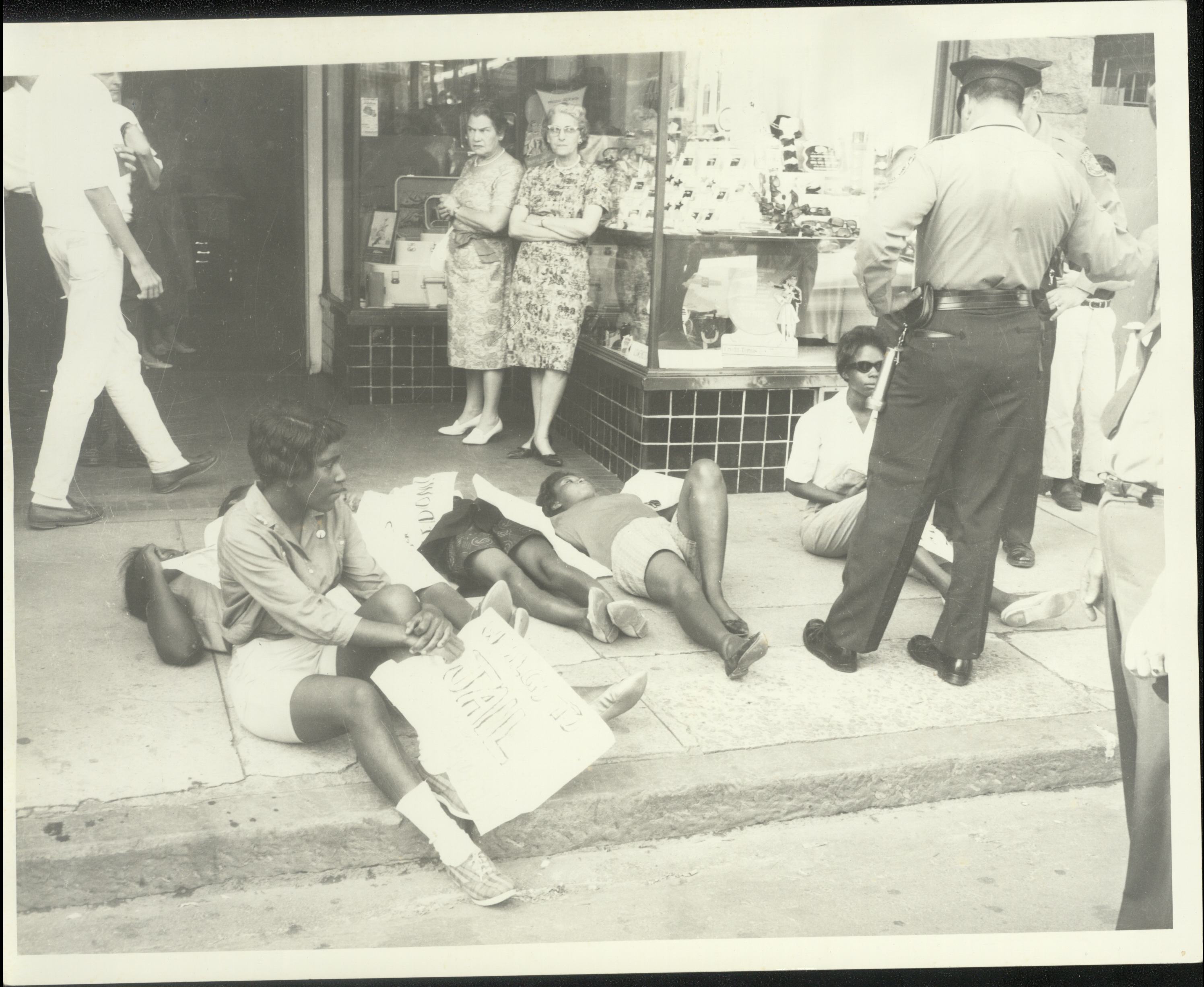 Several demonstrators holding signs lie on the sidewalk outside a Danville business as a police officer and several women stand and watch