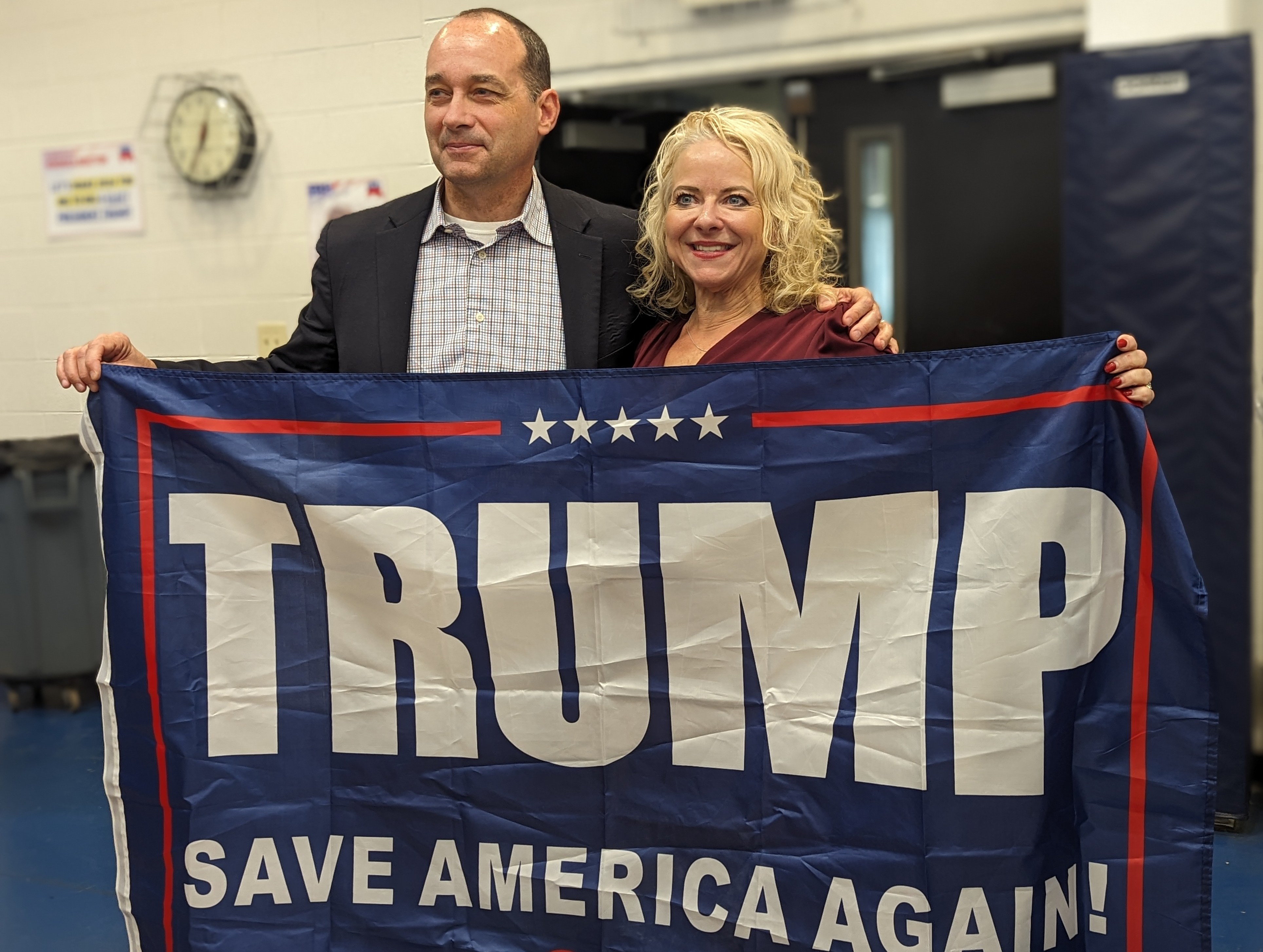 Two politicians stand behind a Trump banner.