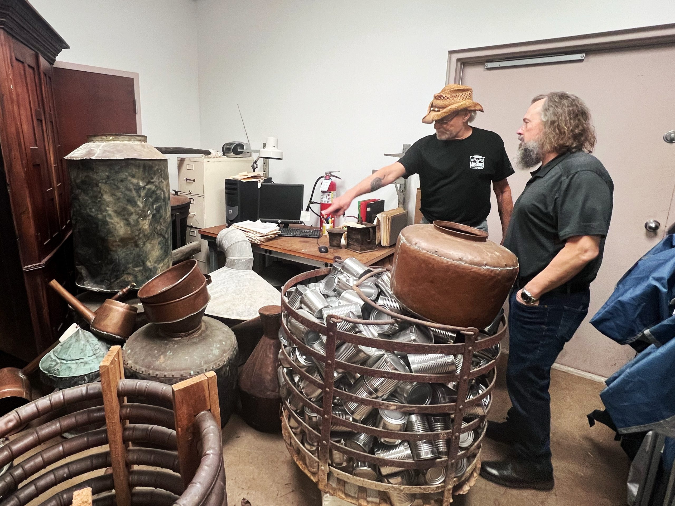 Two men stand in a room filled with old moonshining equipment, including copper kettles and tubing.