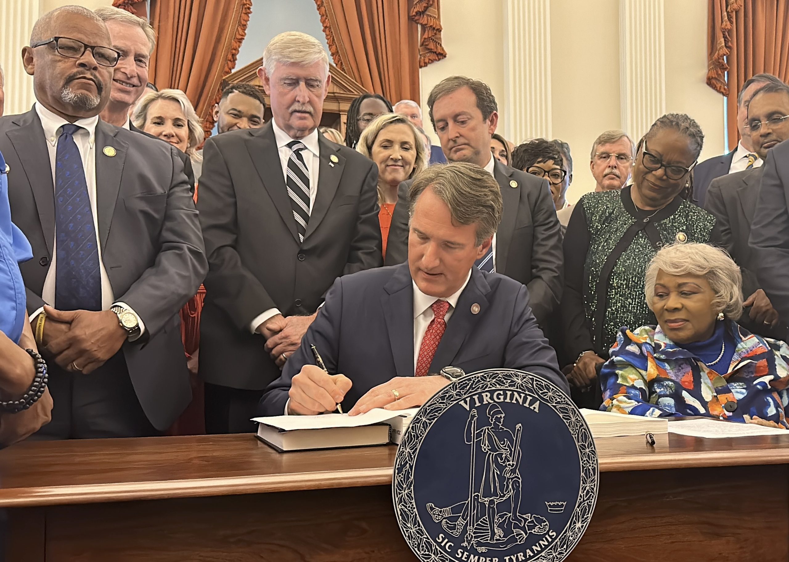 Legislators look on while Gov. Glenn Youngkin signs the state's two-year budget. Photo by Markus Schmidt.