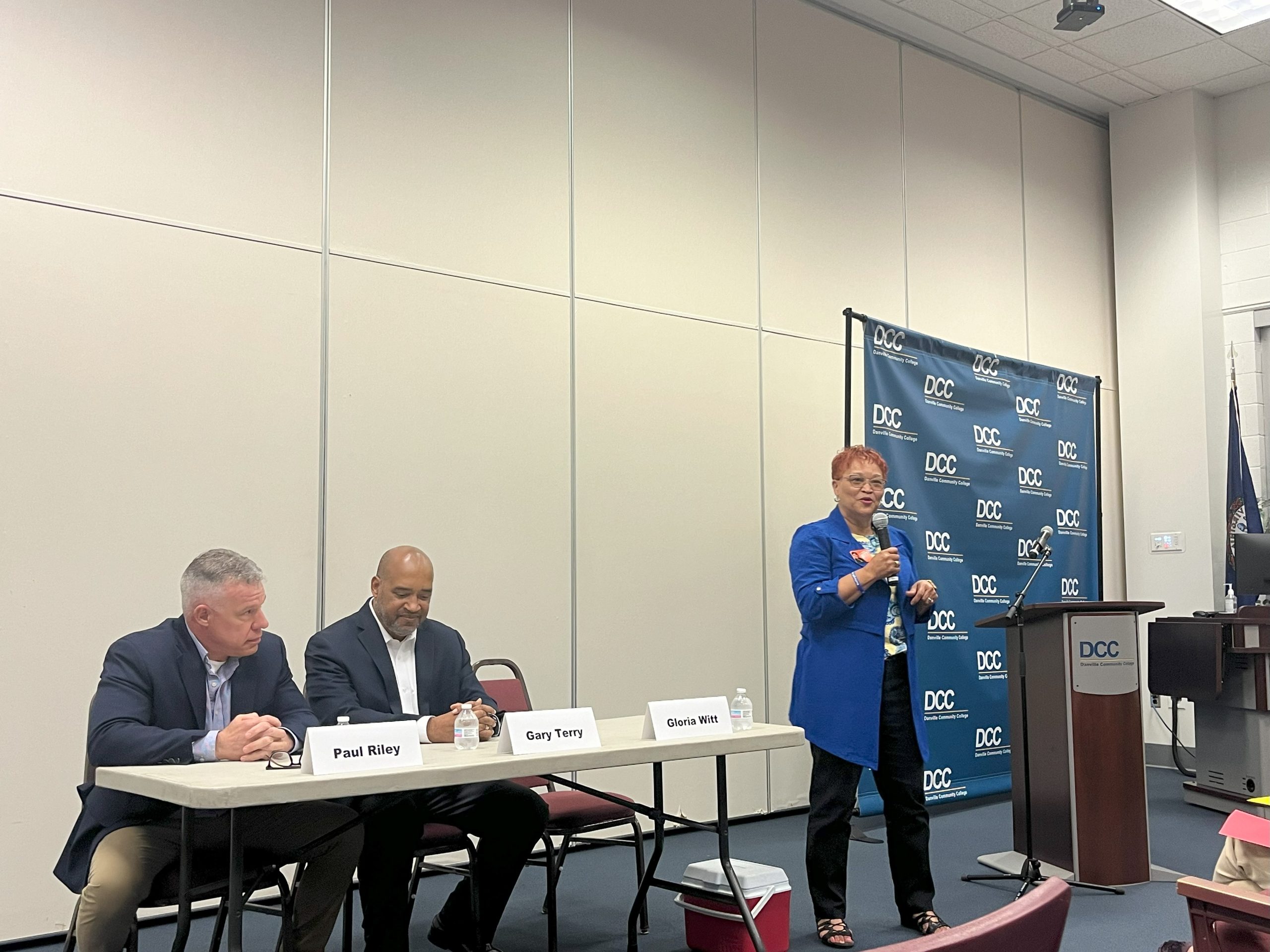 A woman speaks into a microphone, standing next to a table where two men sit as part of a panel of candidates.