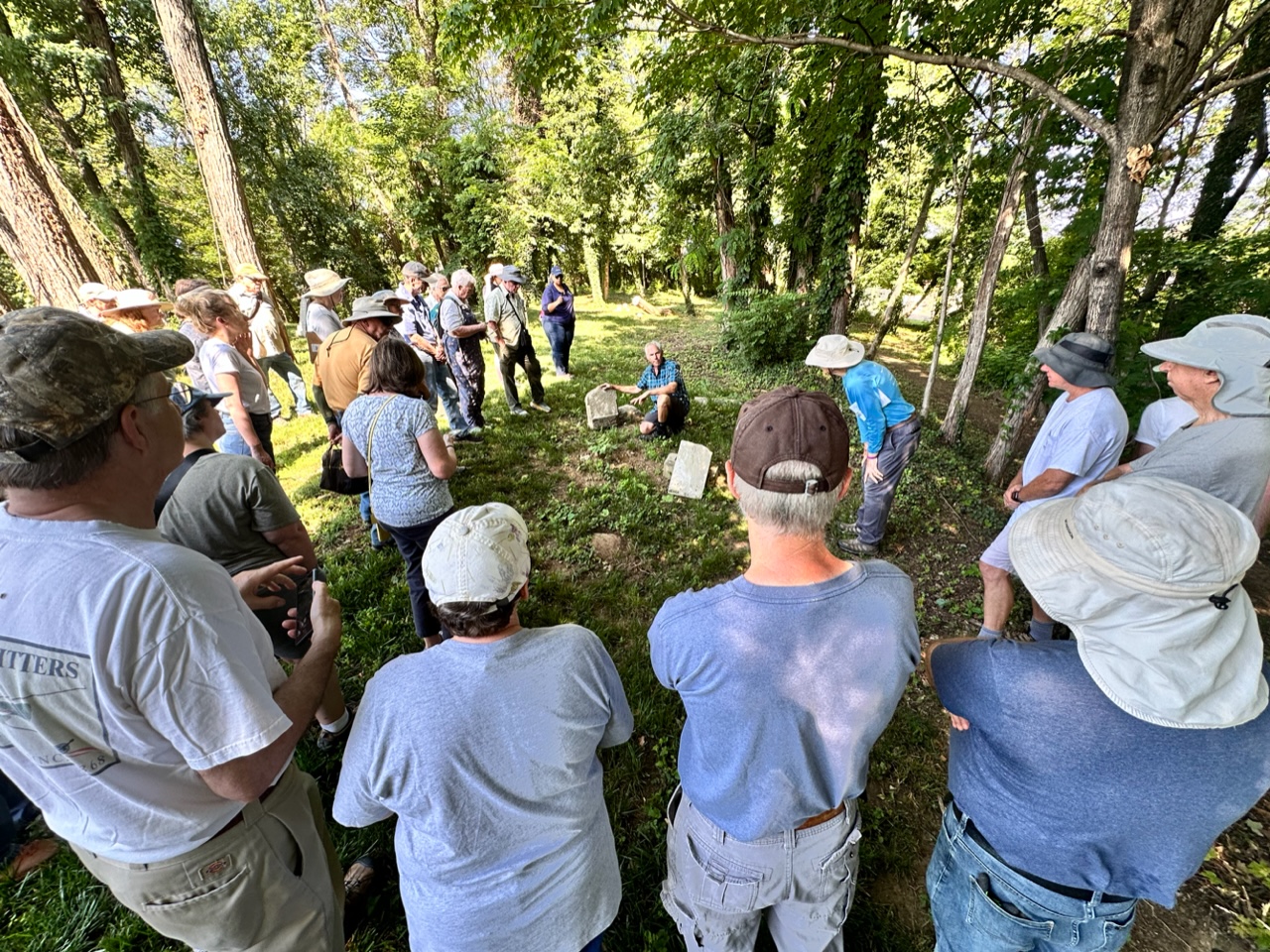Jonathan Appell kneels next to a gravestone in a wooded setting, surrounded by several dozen volunteers.
