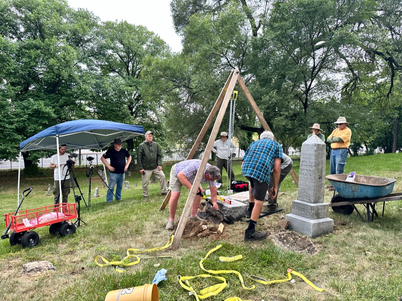A group of people gathered around an old gravestone, using wood beams and other equipment as they work to restore the stone.