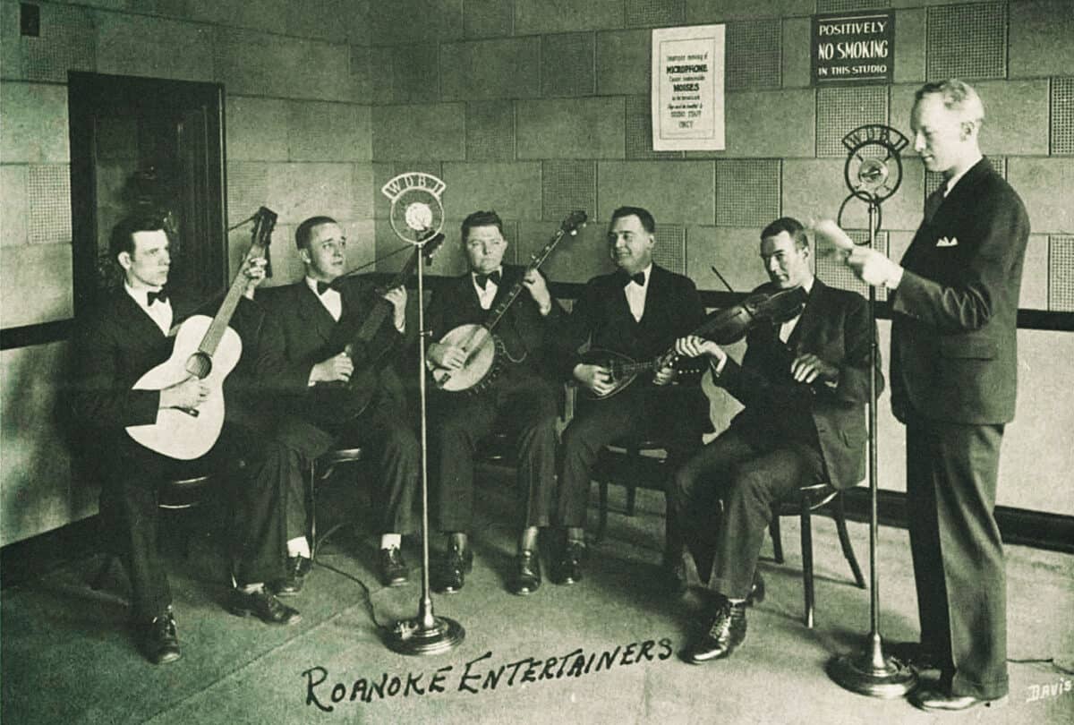 A black-and-white photo of a group of musicians wearing black suits, sitting in an old radio station.