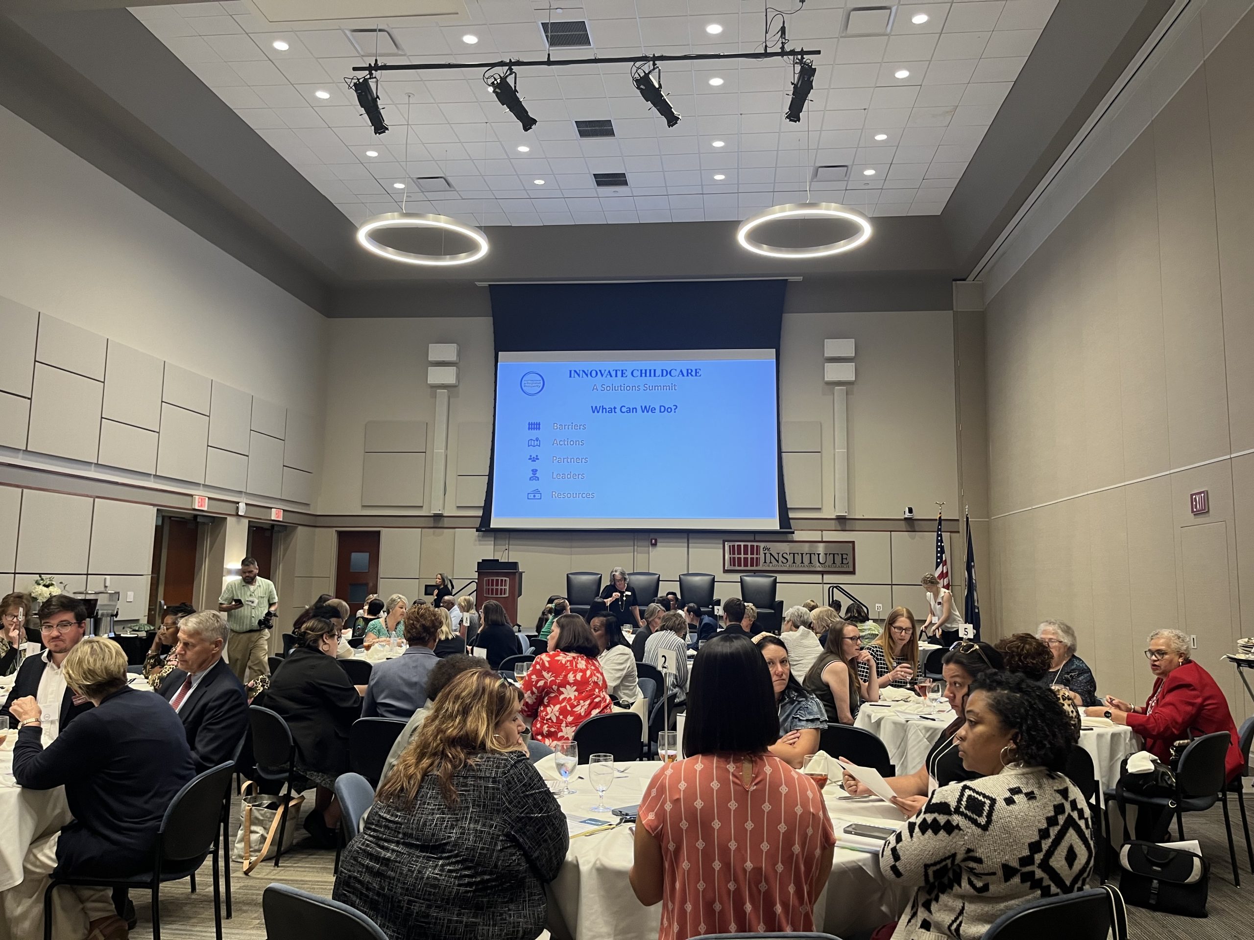 A conference room with people sitting around tables, facing a presentation screen