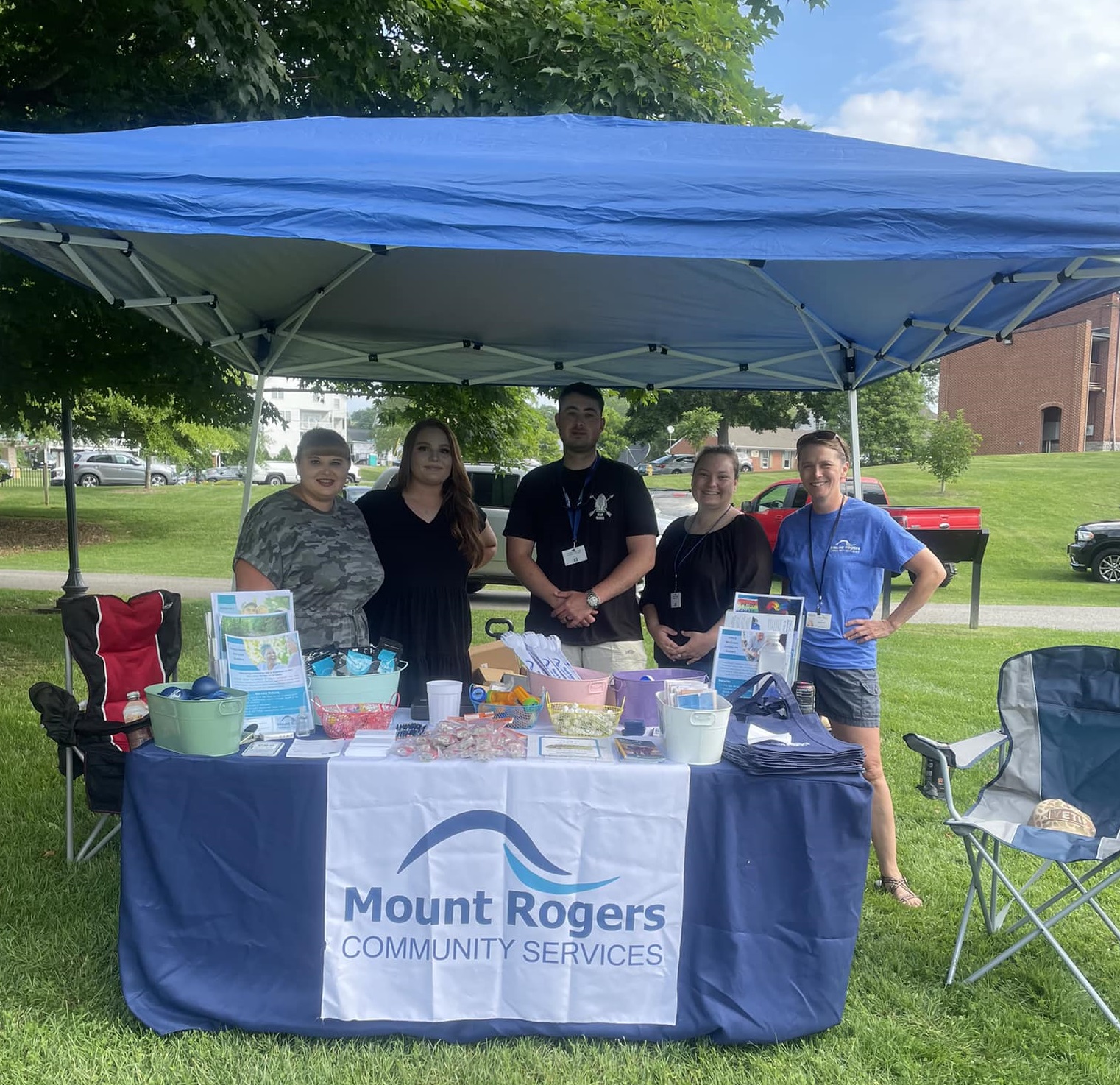 A group of five people under a blue tent in a park, with a "Mount Rogers Community Services" banner.