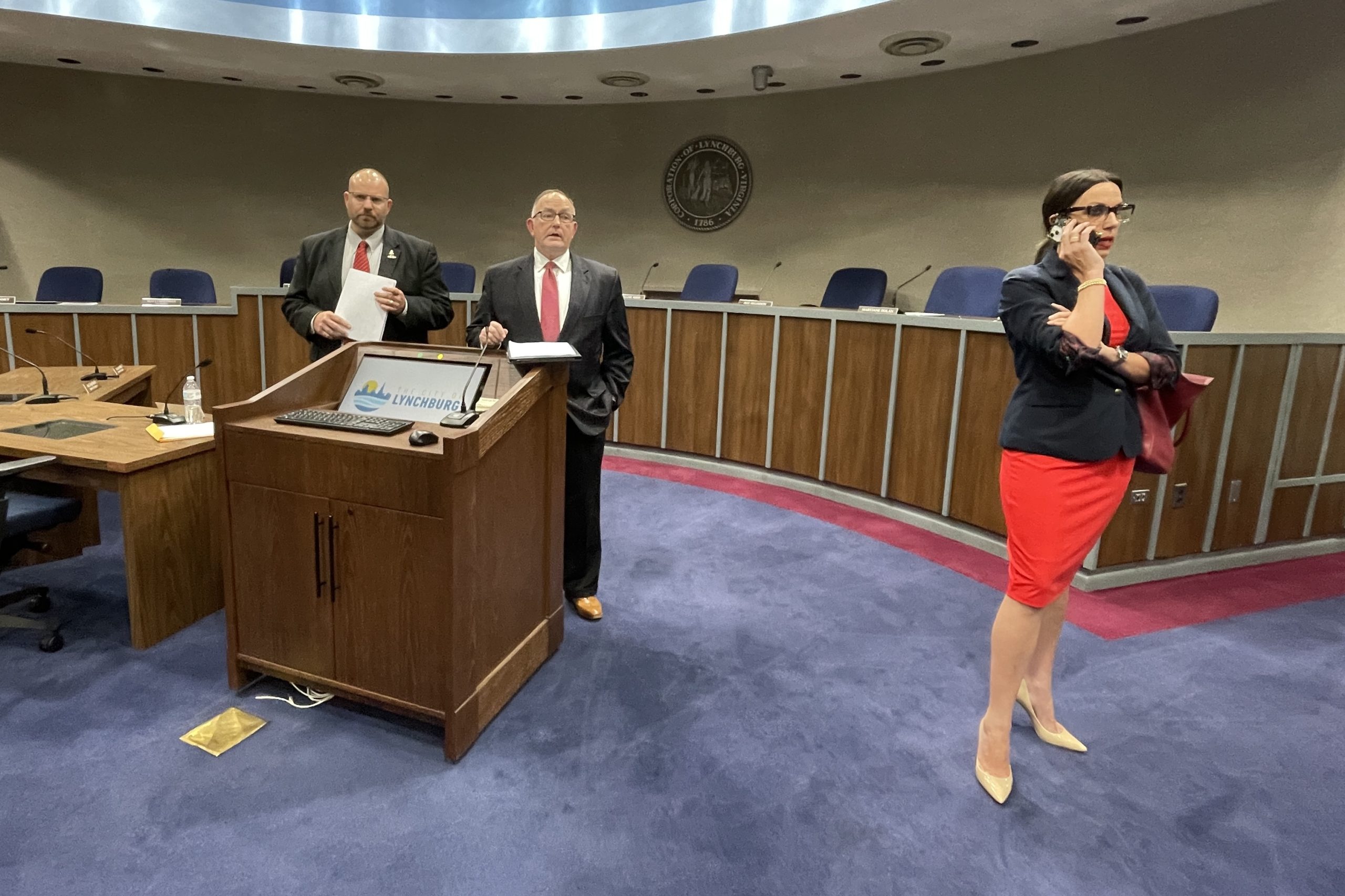 Lynchburg City Council members Marty Misjuns (left) and Jeff Helgeson (center) hold a press conference in council chambers in Lynchburg City Hall as Mayor Stephanie Reed (right) speaks on the phone as she attempts to make the news media leave the room.