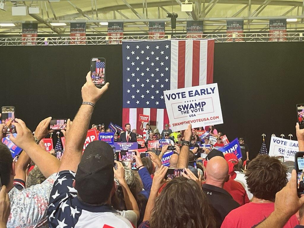 JD Vance in the background, in front of a huge American flag, with a big crowd standing in front of him.
