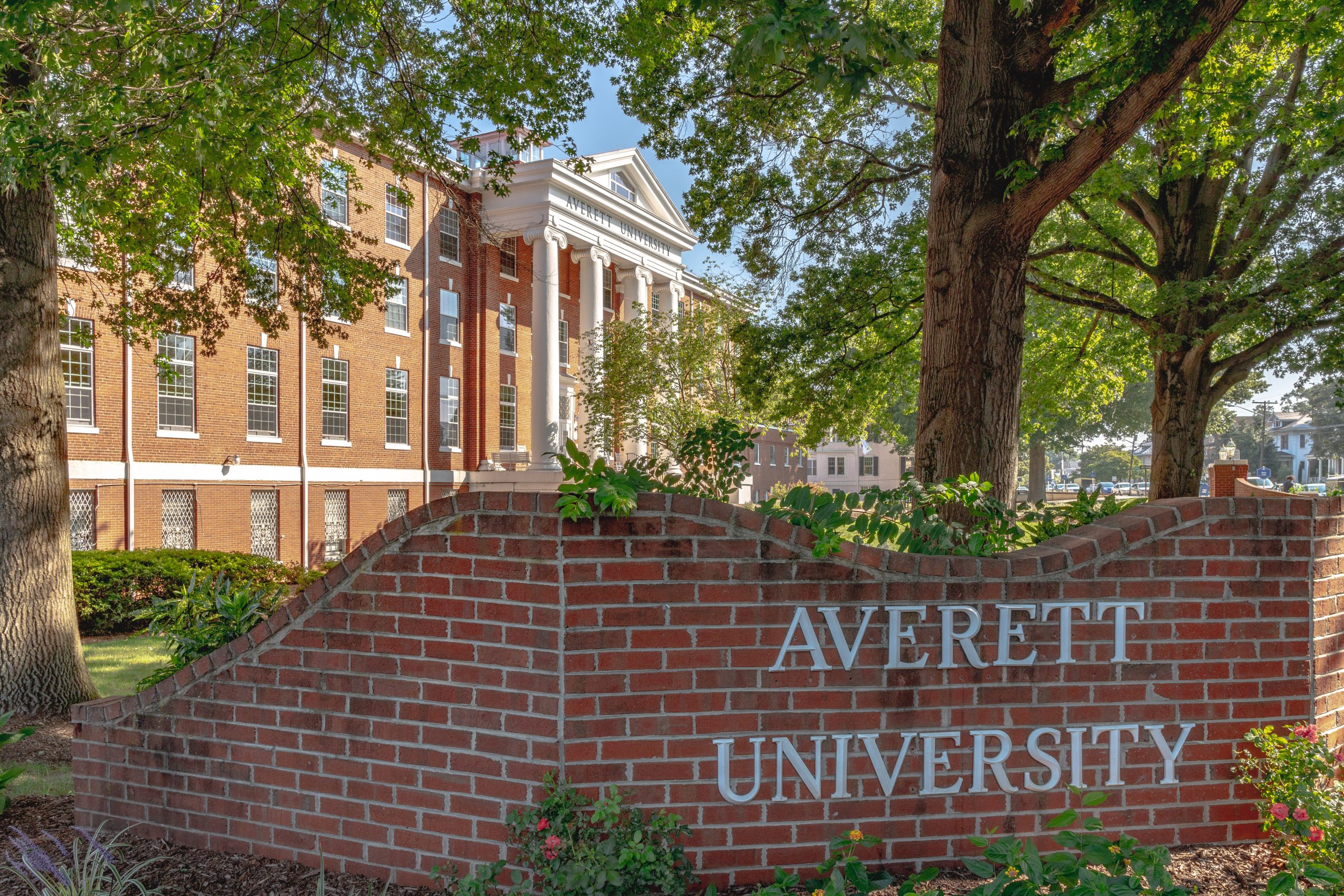 A red brick sign saying "Averett University" in the foreground, with a large red-brick building with white columns in the background.