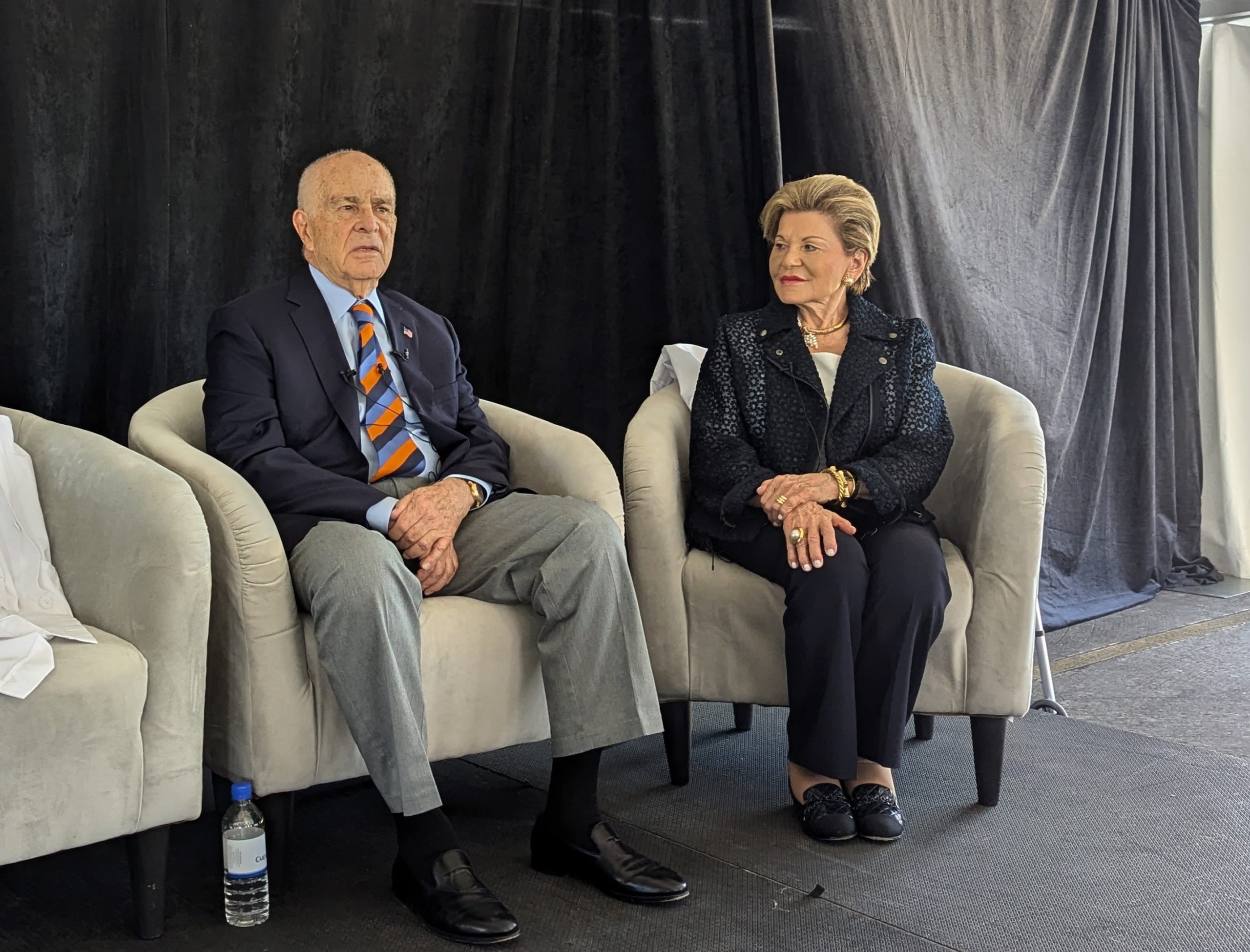 Nick and Jenny Taubman sit in upholstered chairs on a stage with a black backdrop.