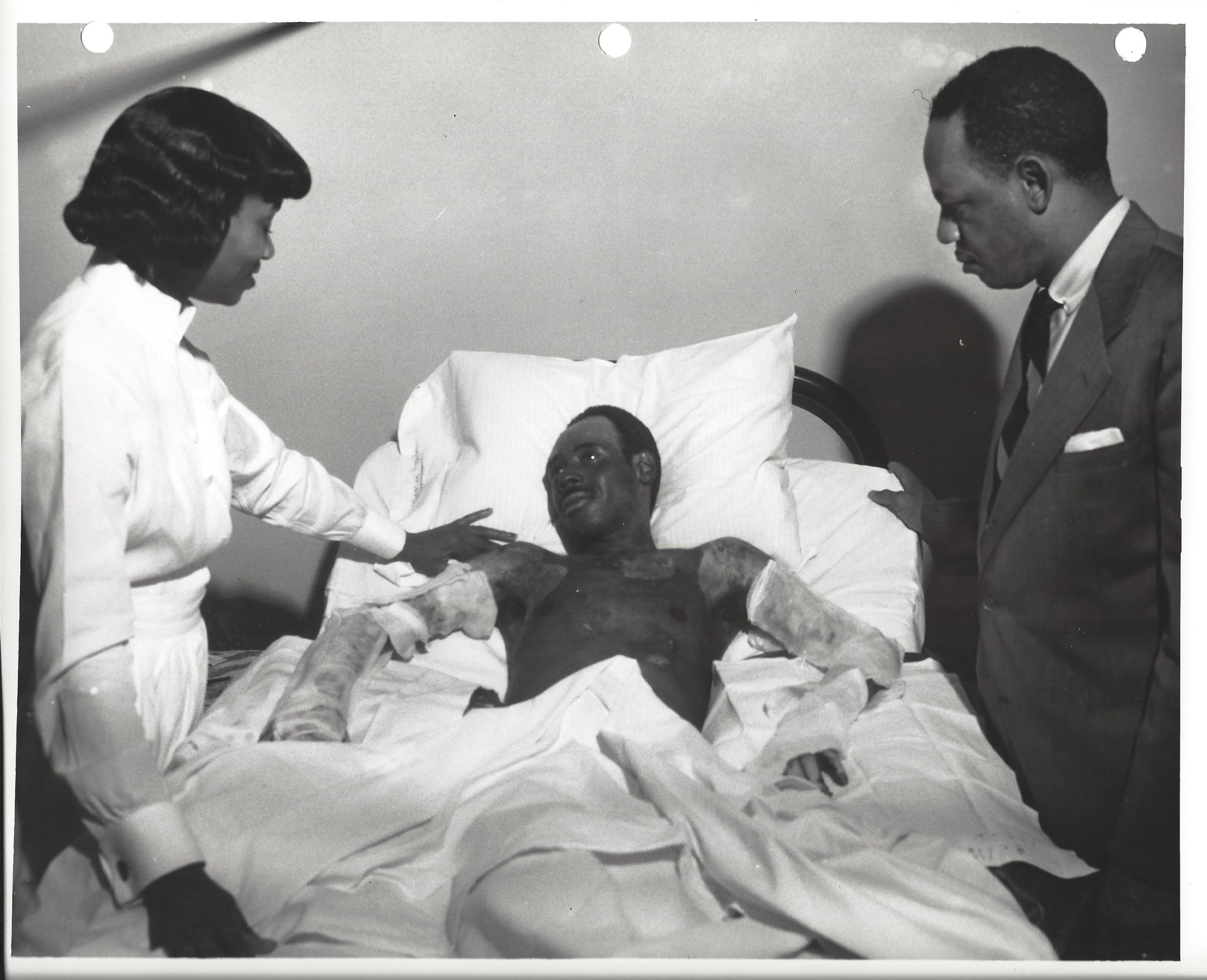 A man in a suit and a woman in a nurse's uniform stand over a hospital bed with a patient at Winslow Hospital.