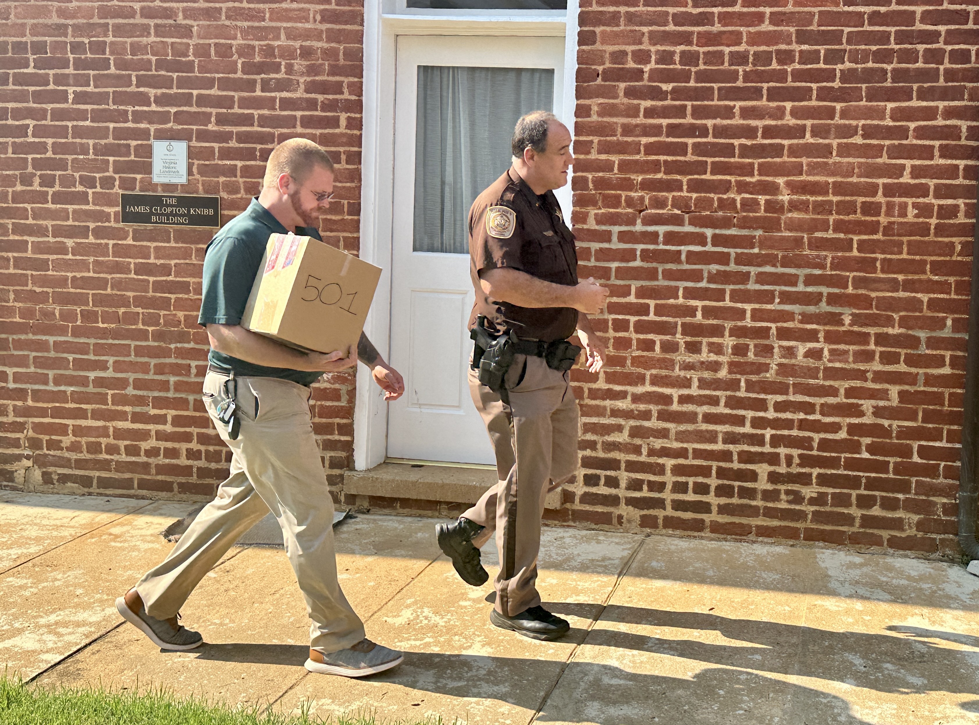 A man carrying a cardboard box walks with a sheriff's deputy ouside a red brick building in Goochland County