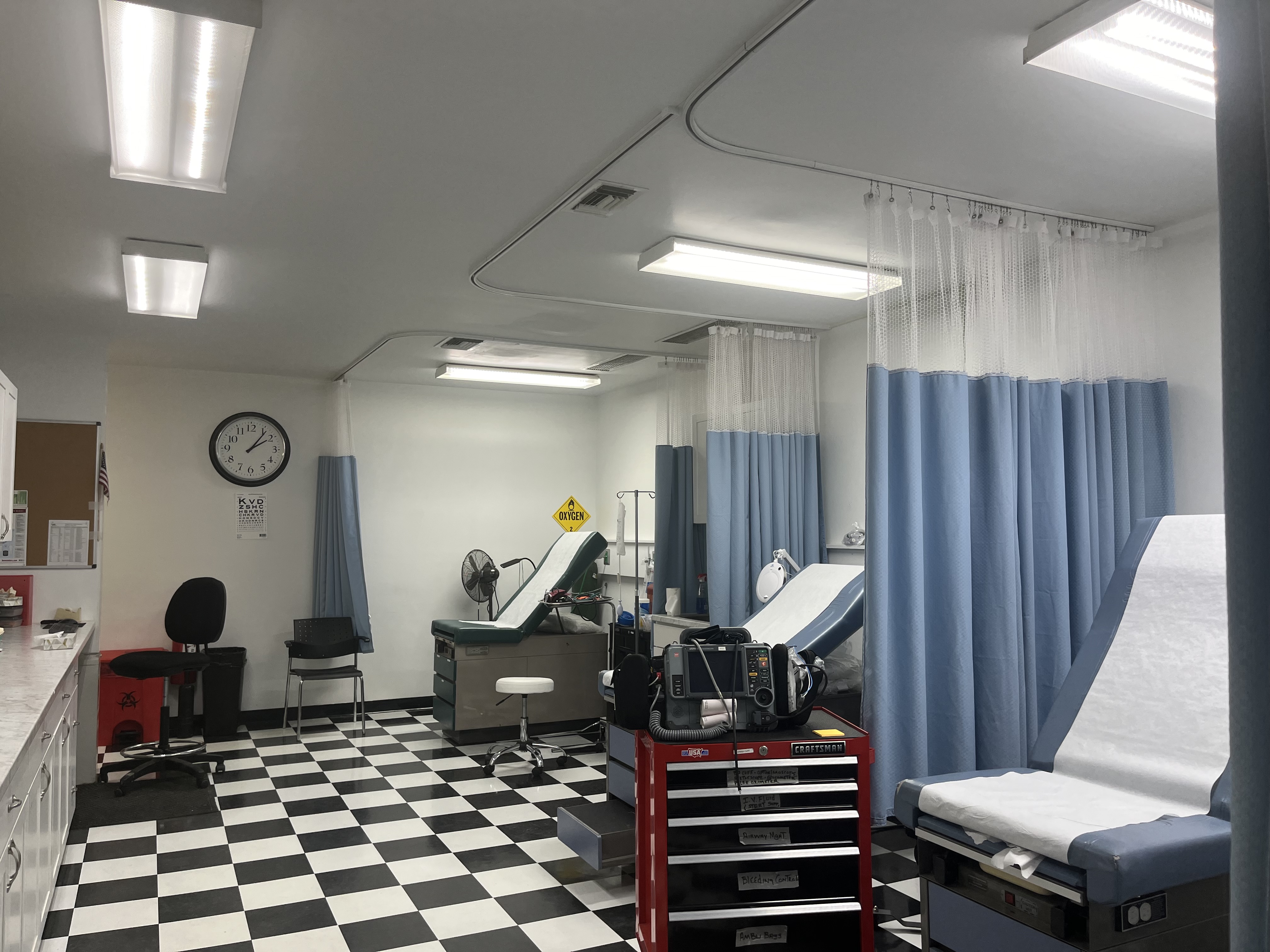 A small clinic, which looks like a room in a doctors office, has a curtain separating chairs for patients and other medical supplies. 