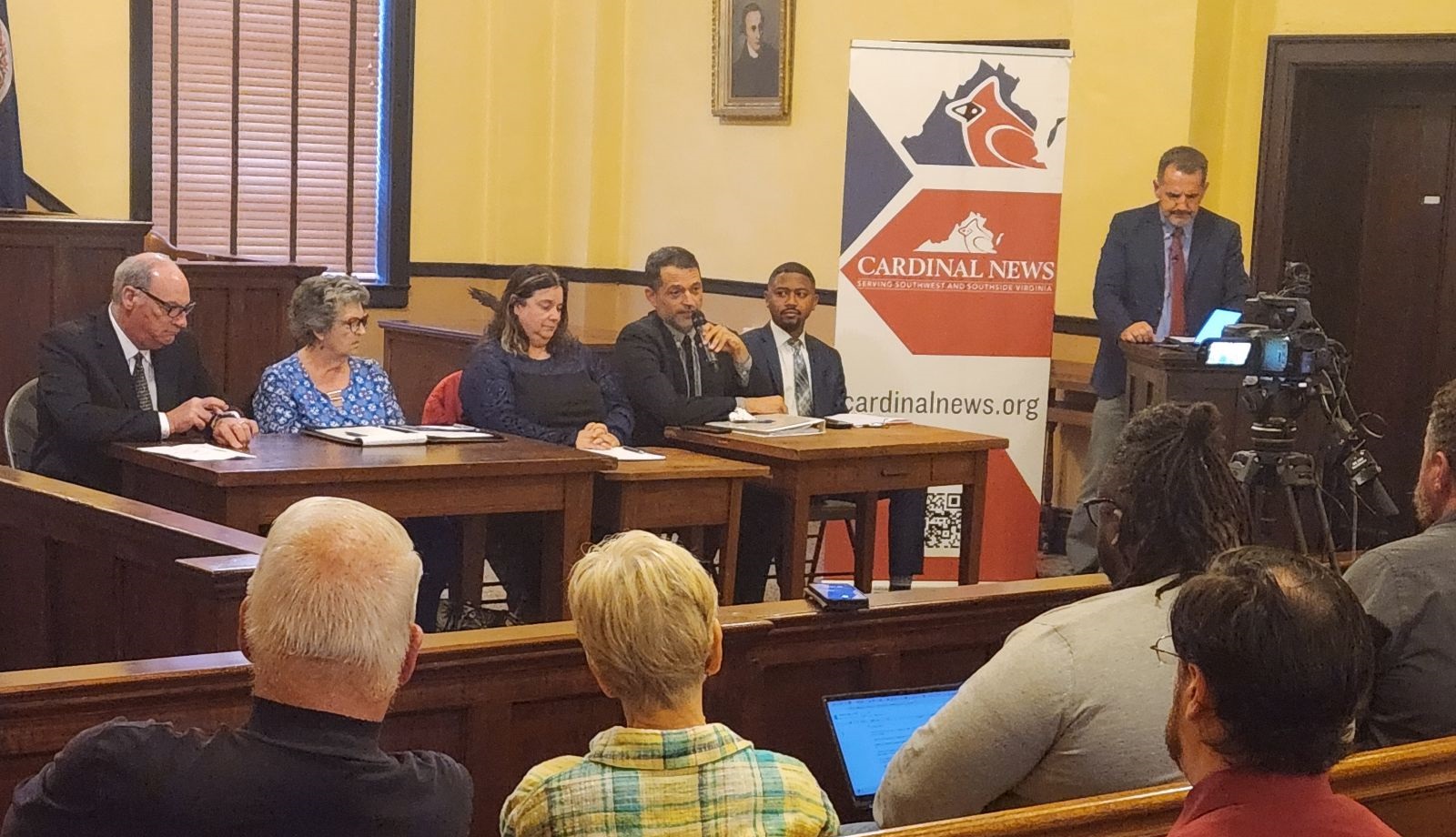 Martinsville City Council candidates (from left) John Wilson, Kathy Lawson, LaNita Herlem, Julian Mei and Rayshaun Graveley sit at a table in front of a room, while Terry Young of Patrick and Henry Community College stands to their left at a lectern.