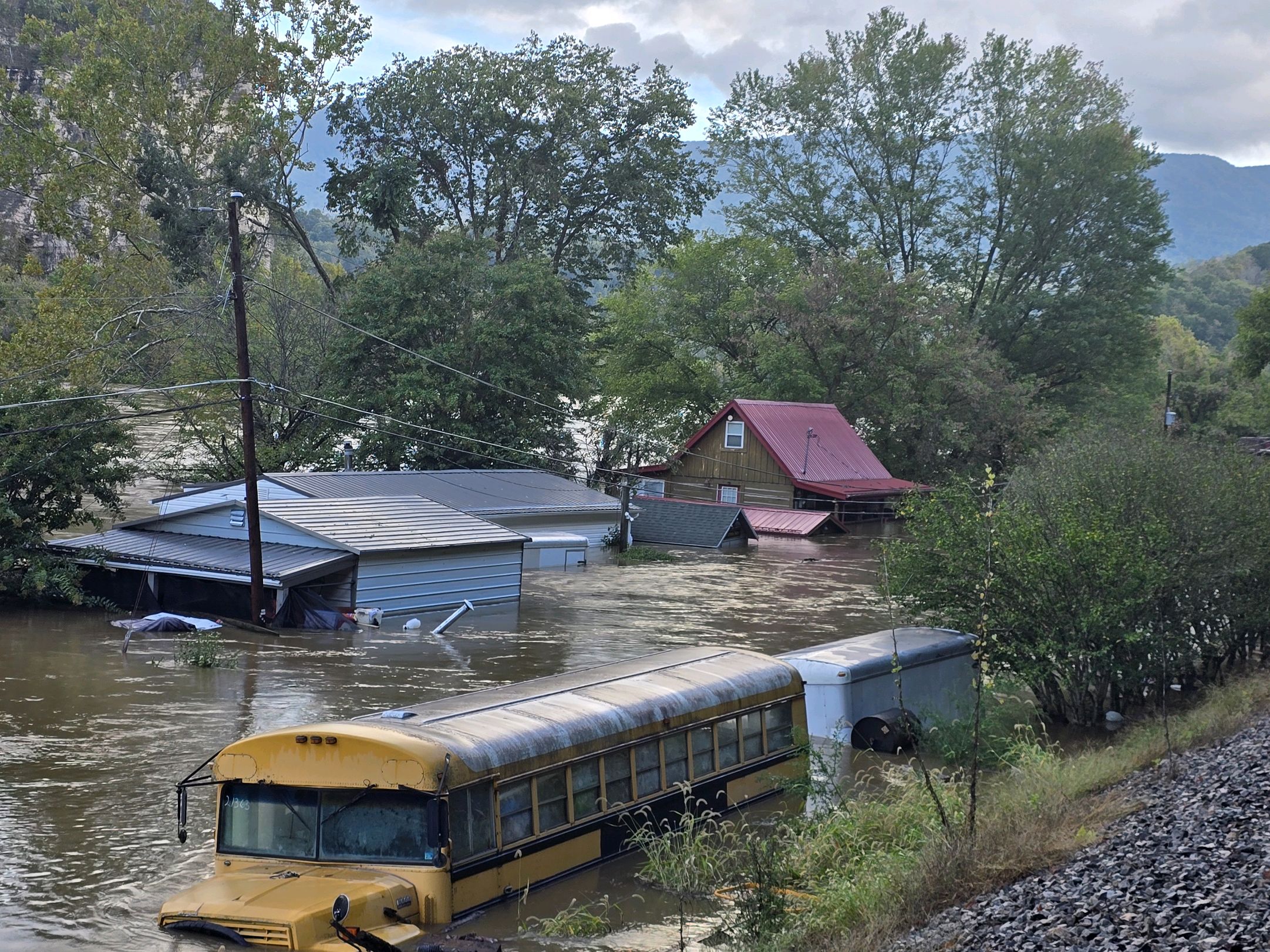 A house, several outbuildings and a school bus surrounded by muddy brown floodwaters.