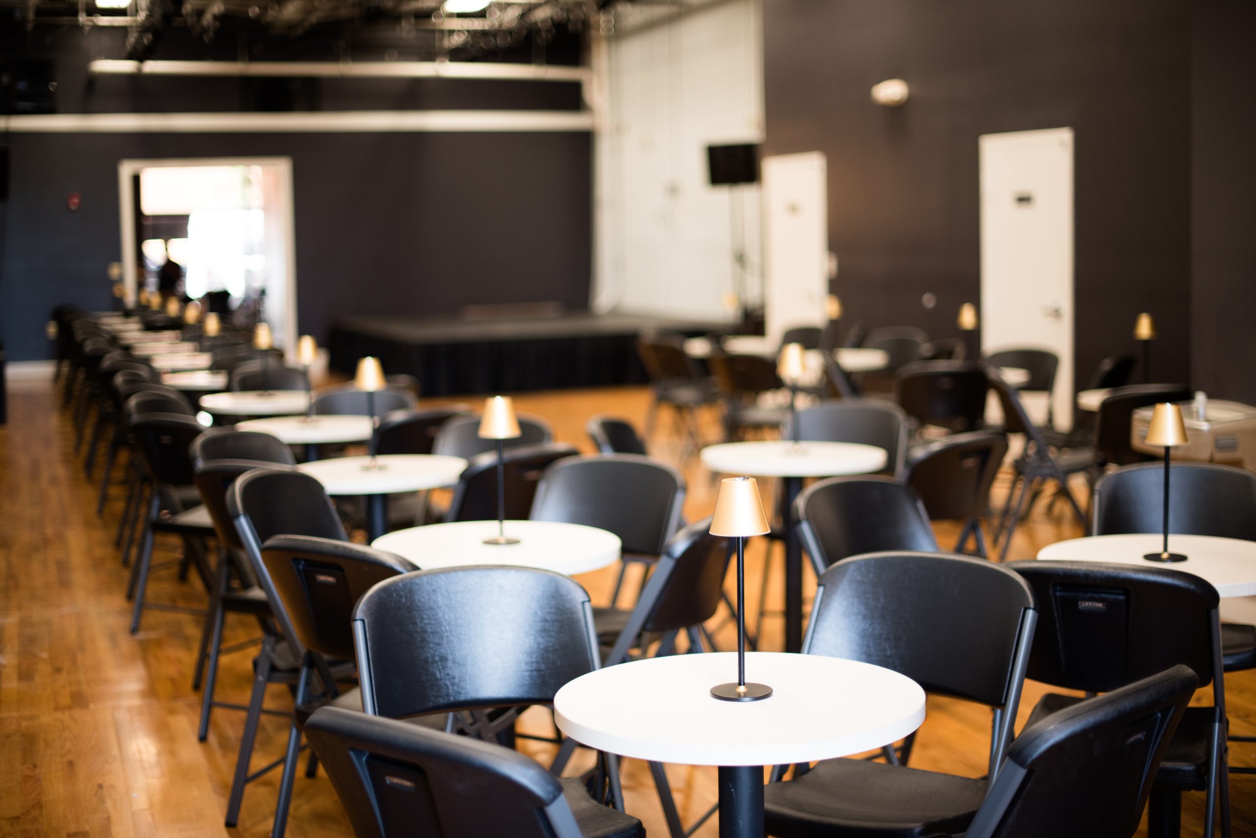 A small music venue, with 4-top tables and black chairs in several rows leading to a small stage in the corner.
