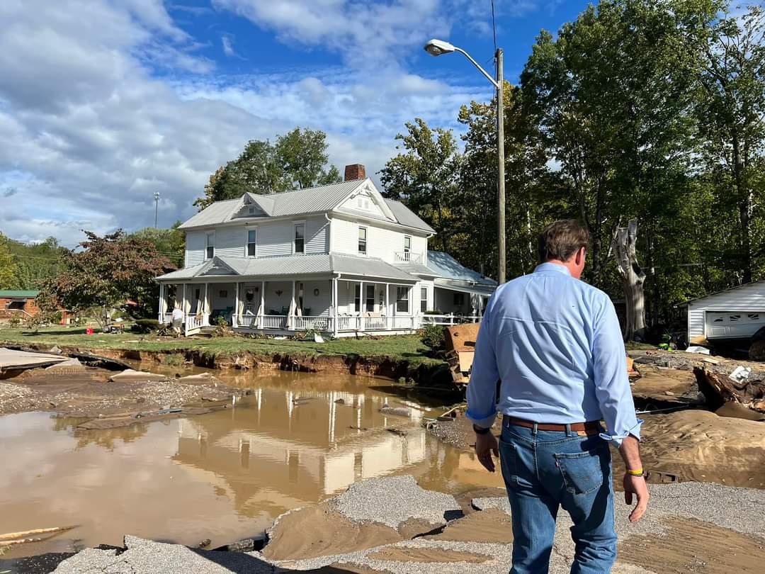 Gov. Glenn Youngkin walks down a flooded street in Damascus, with a large white house in the background.