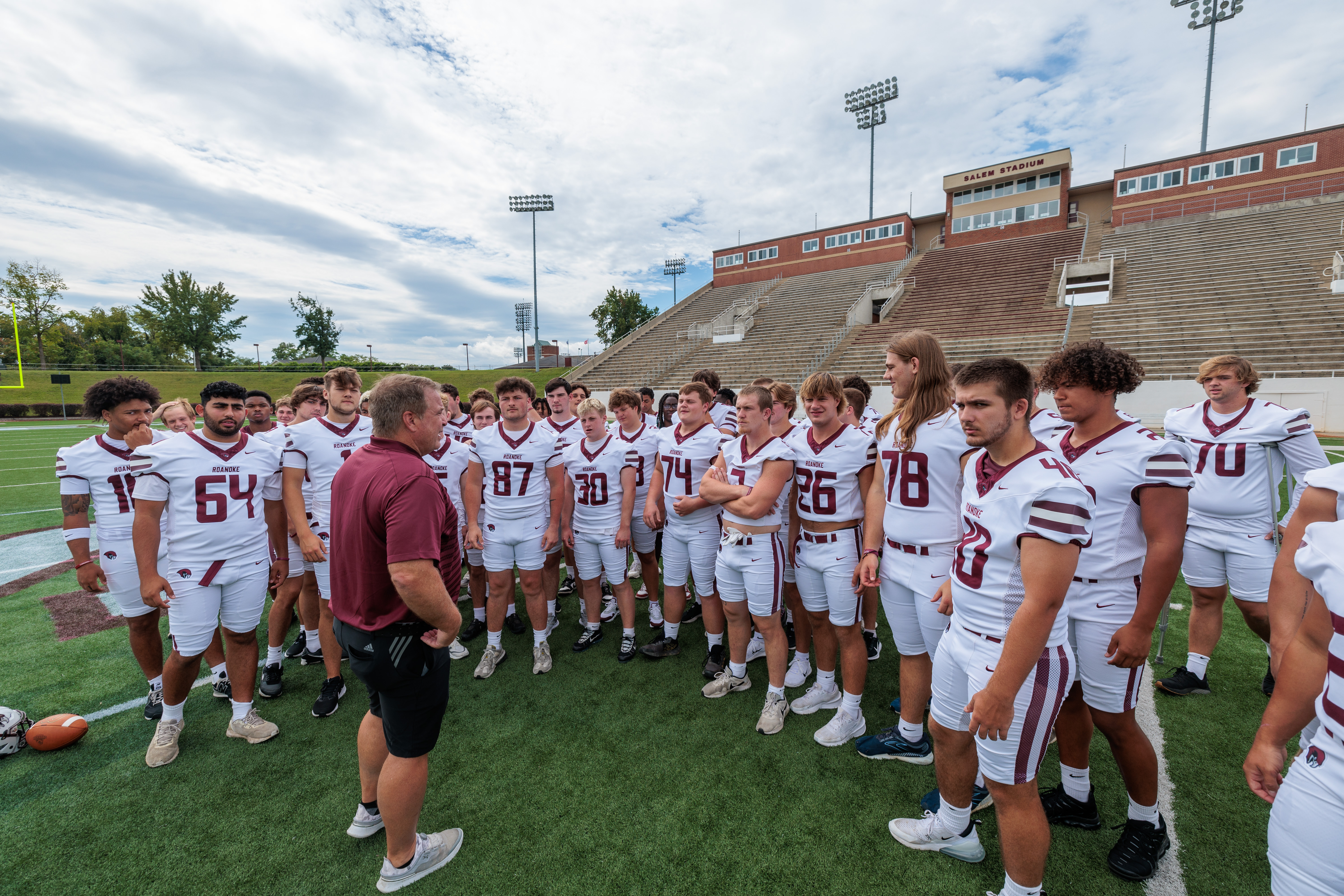 Coach Bryan Stinespring talks to the Roanoke College football players who are gathered around him on the field in a semi-circle, wearing white uniforms.