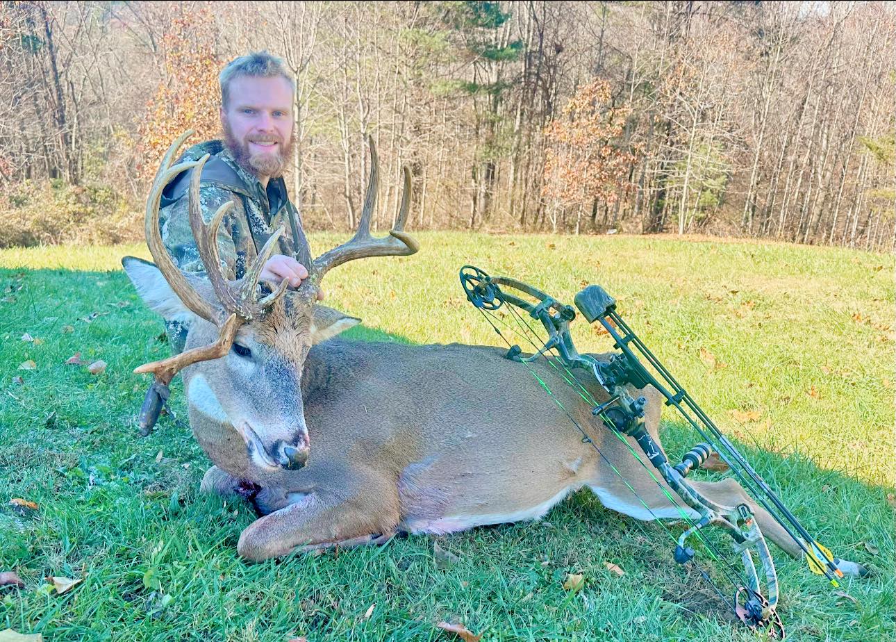 A man in hunting clothes kneels down next to a dead buck in a field.