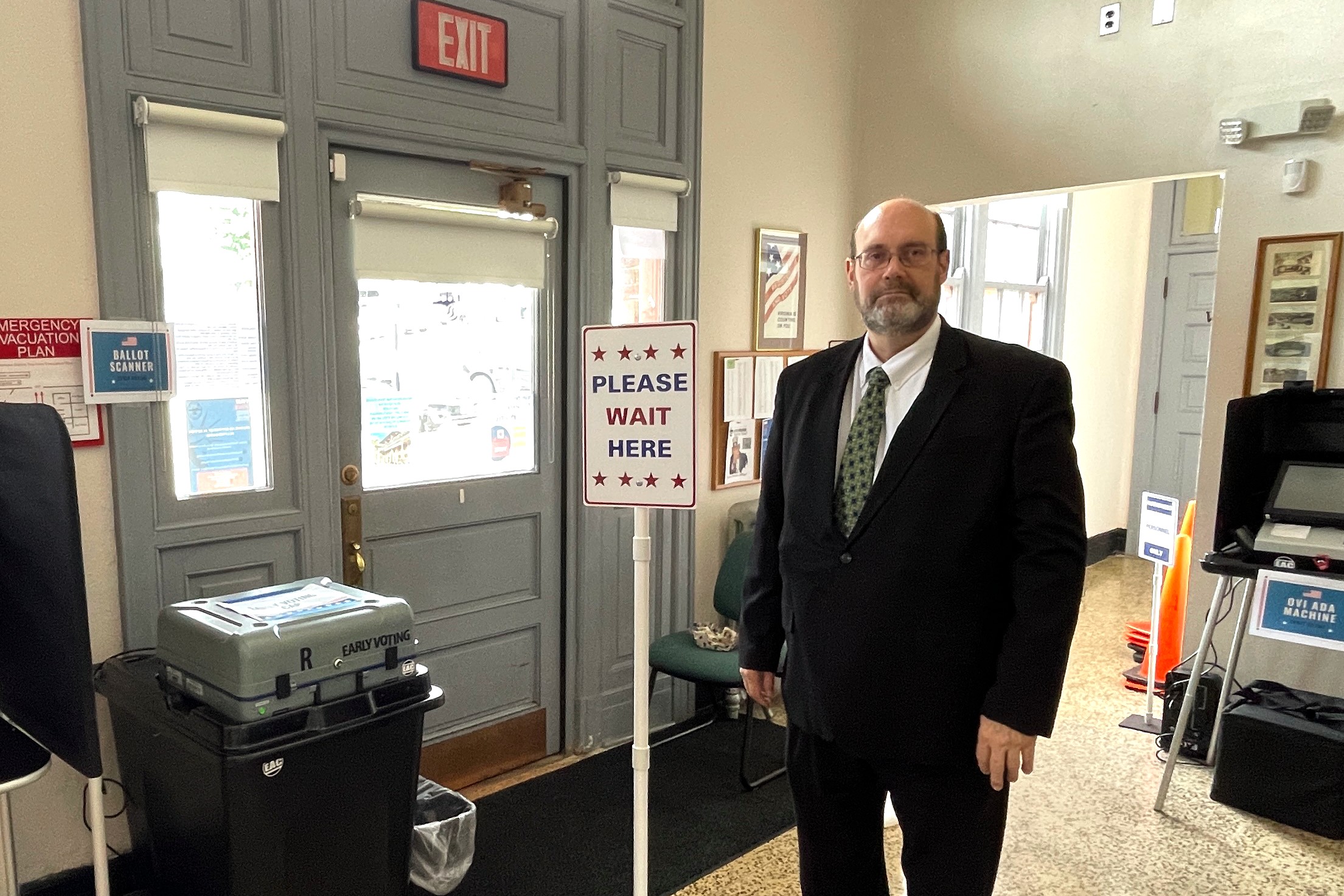 A man in a suit stands by a ballot scanner machine and a sign that reads "Please wait here."