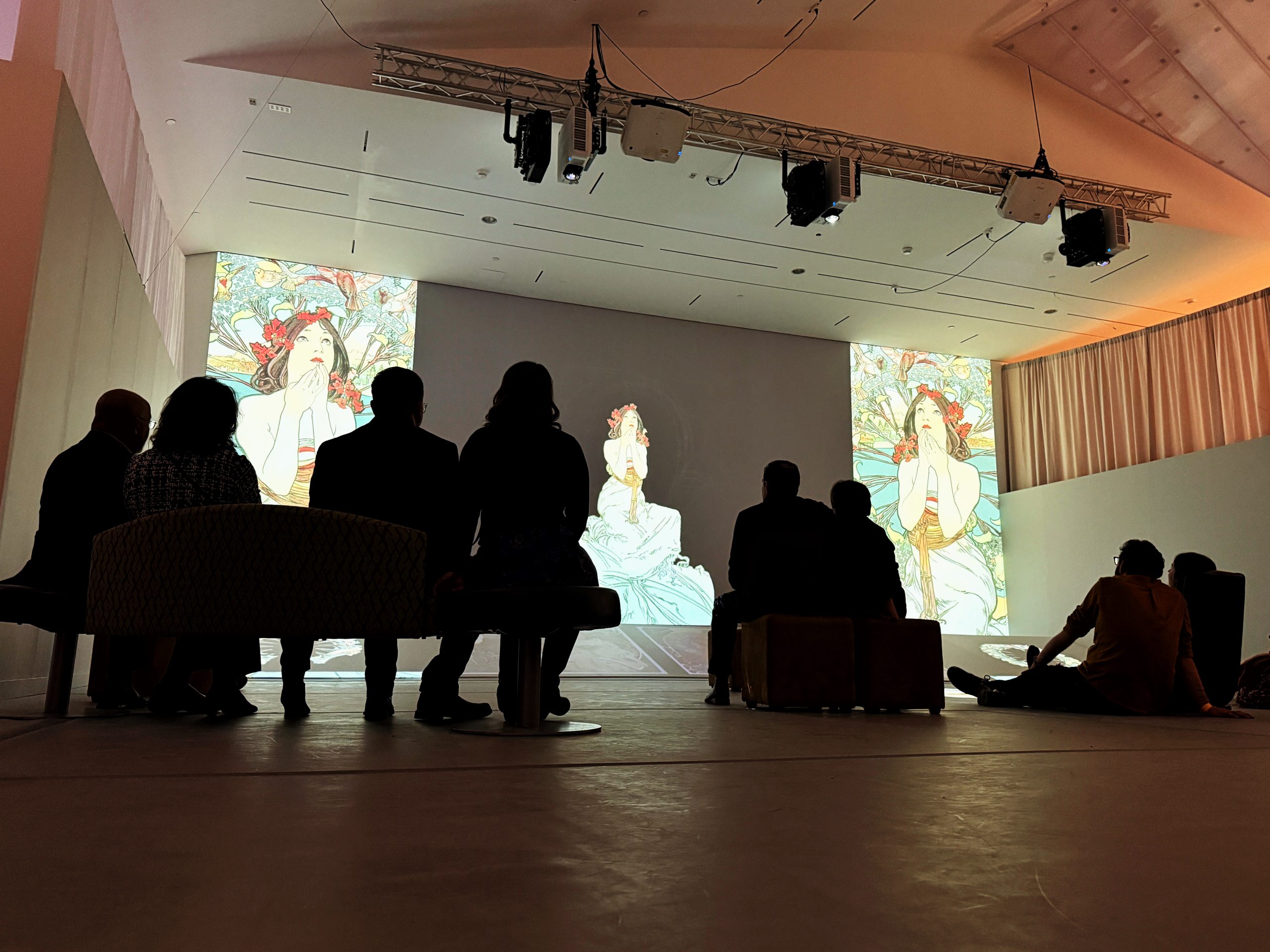 Museum visitors sit in the shadows as Art Nouvea images of a woman gazing thoughtfully skyward are projected on the wall. Photo by Lindsey Hull.