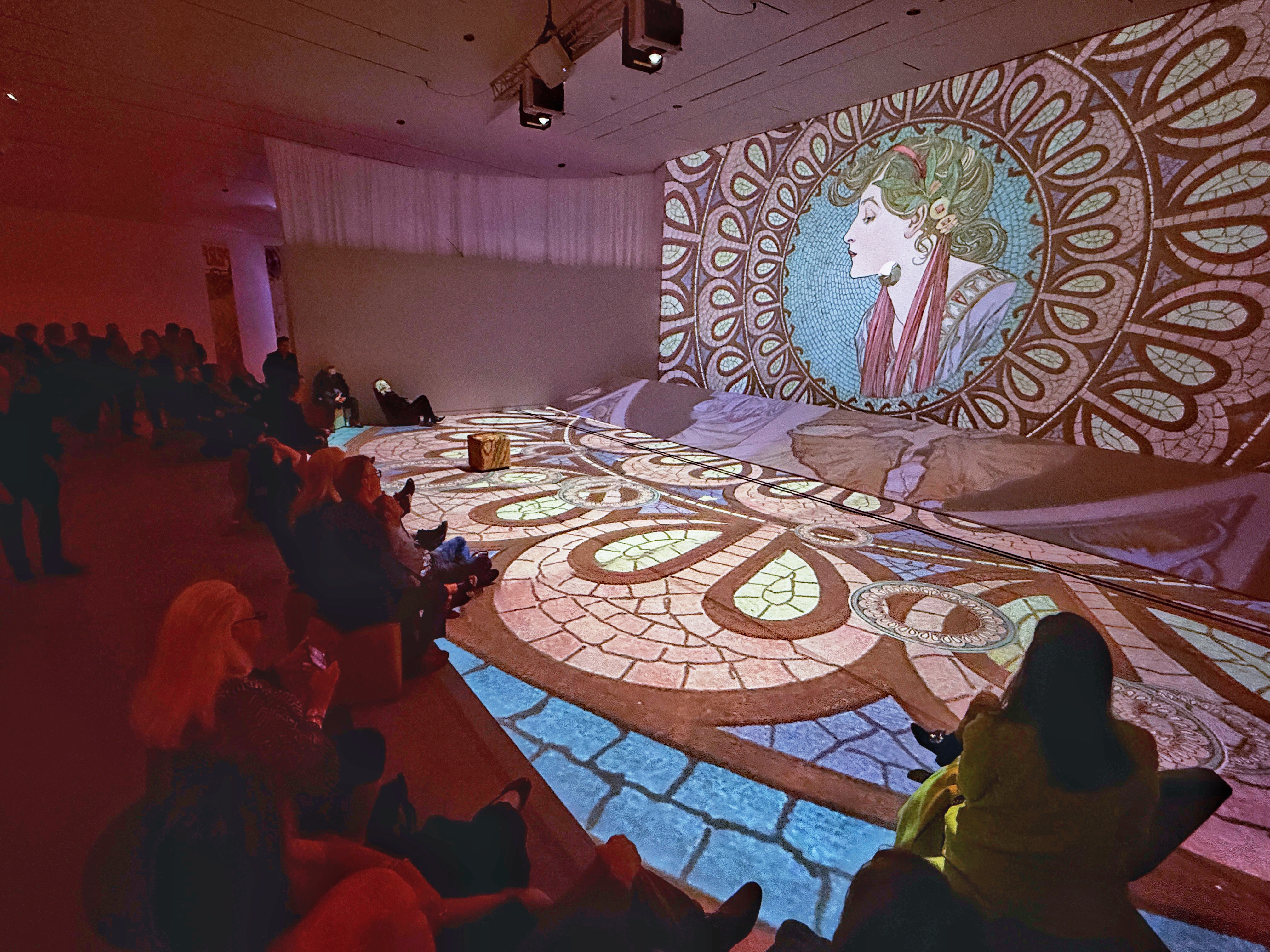 People in shadow watch a large projection of an Art Nouveau graphic design of a woman with leaves and flowers in her hair.