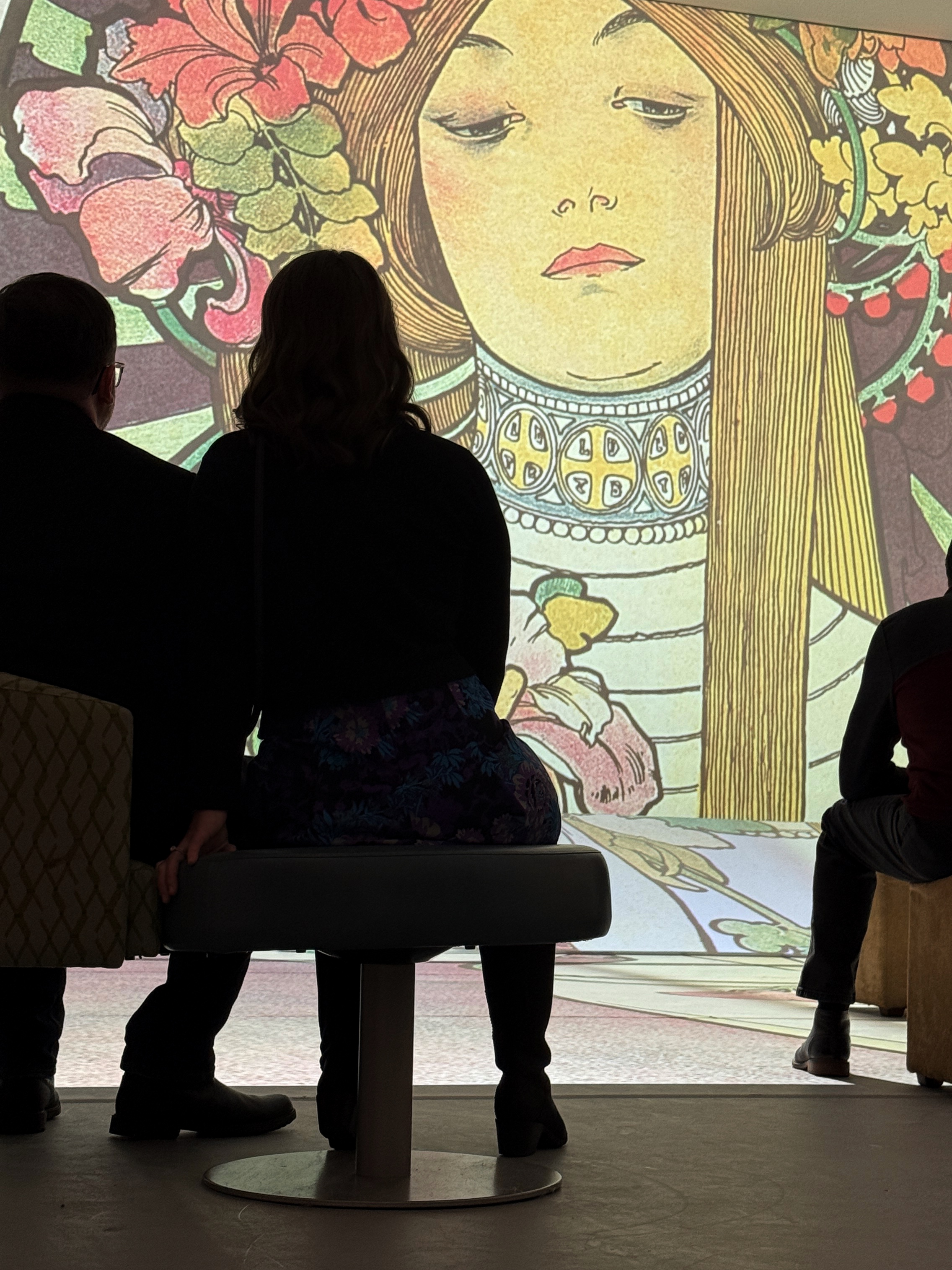 Visitors sit in shadow as 18 foot image of Art Nouveau woman, her hair adorned in flowers, is projected on the wall in front of them.