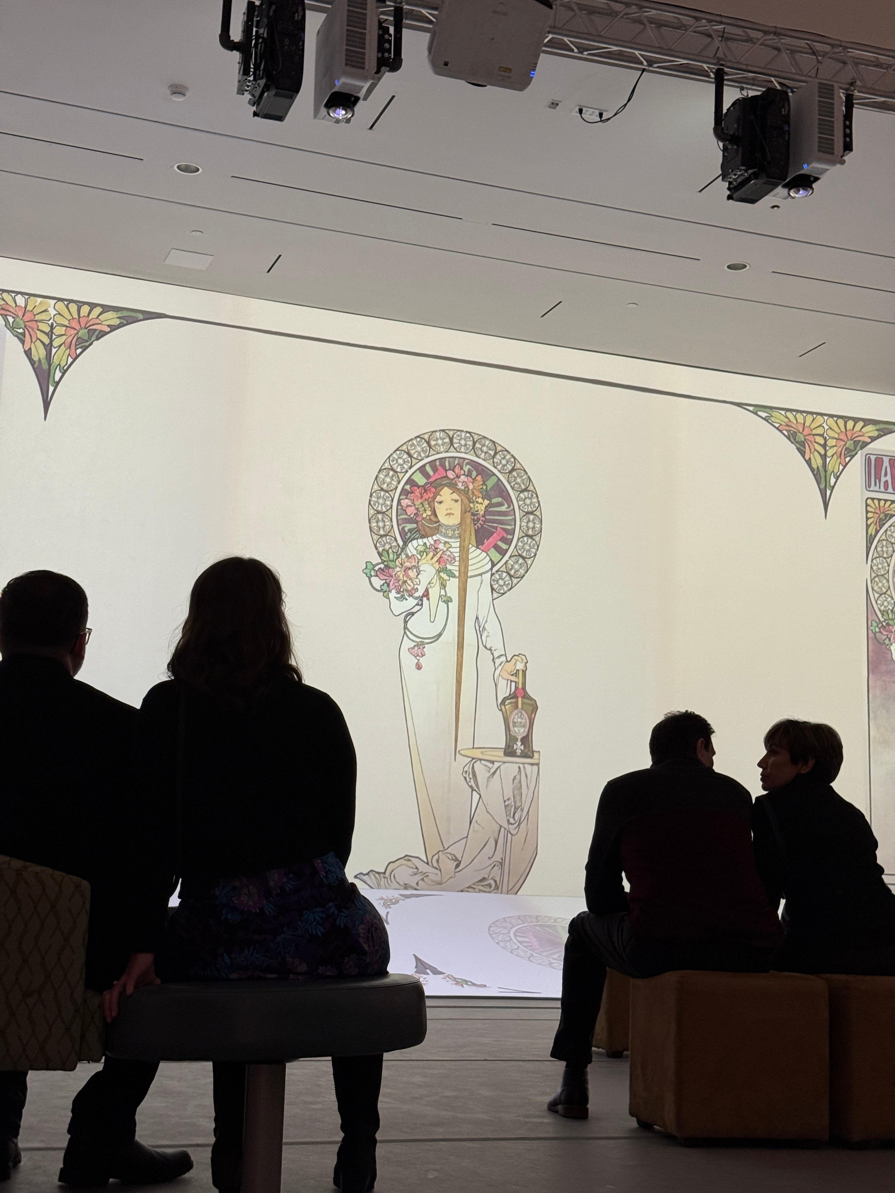 Two couples sit on benches and seats in the shadows as an Art Nouveau image is projected on a wall beyond them.