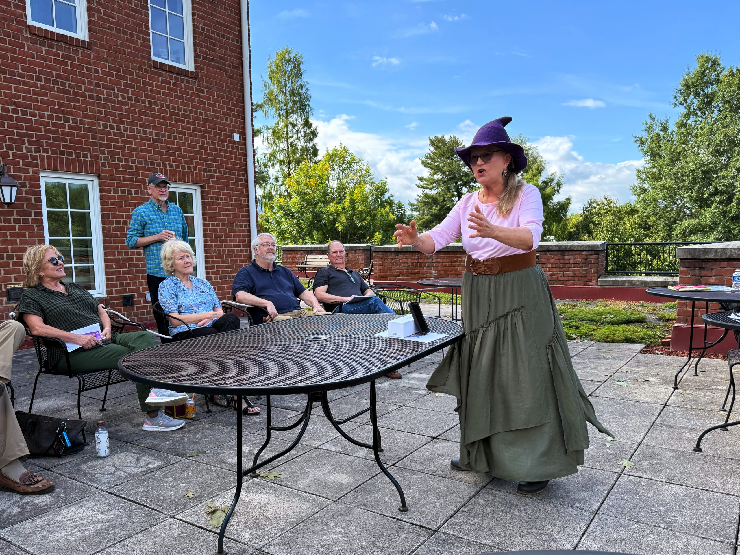 A woman wearing a witches hat stands on a patio with a group of people seated on chairs around her.