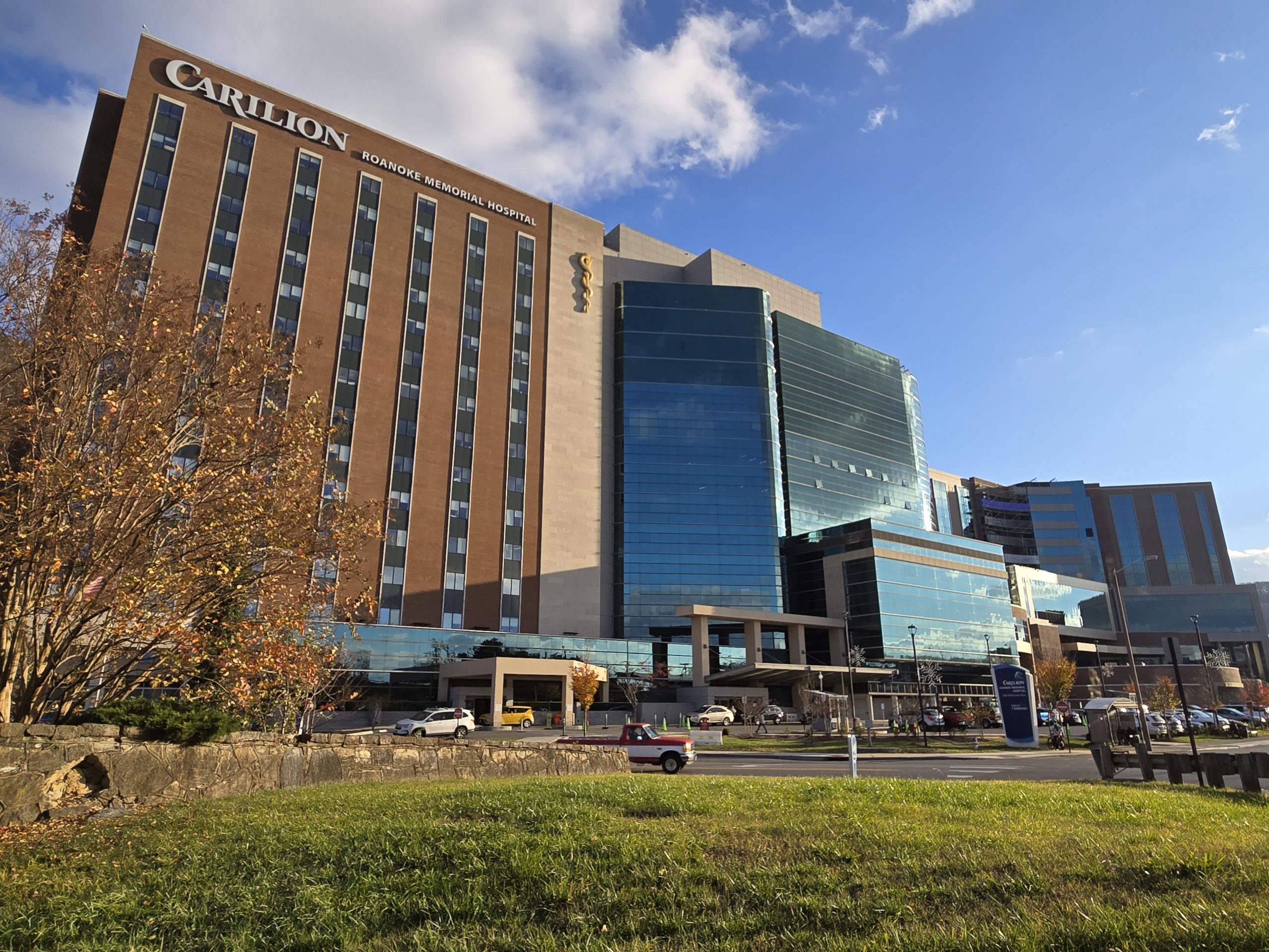 Carilion Roanoke Memorial Hospital on a November day. The building is a mix modern architecture with large glass windows and a taller brick structure. Autumn foliage with golden leaves is visible in the foreground, contrasting with the hospital's sleek design.