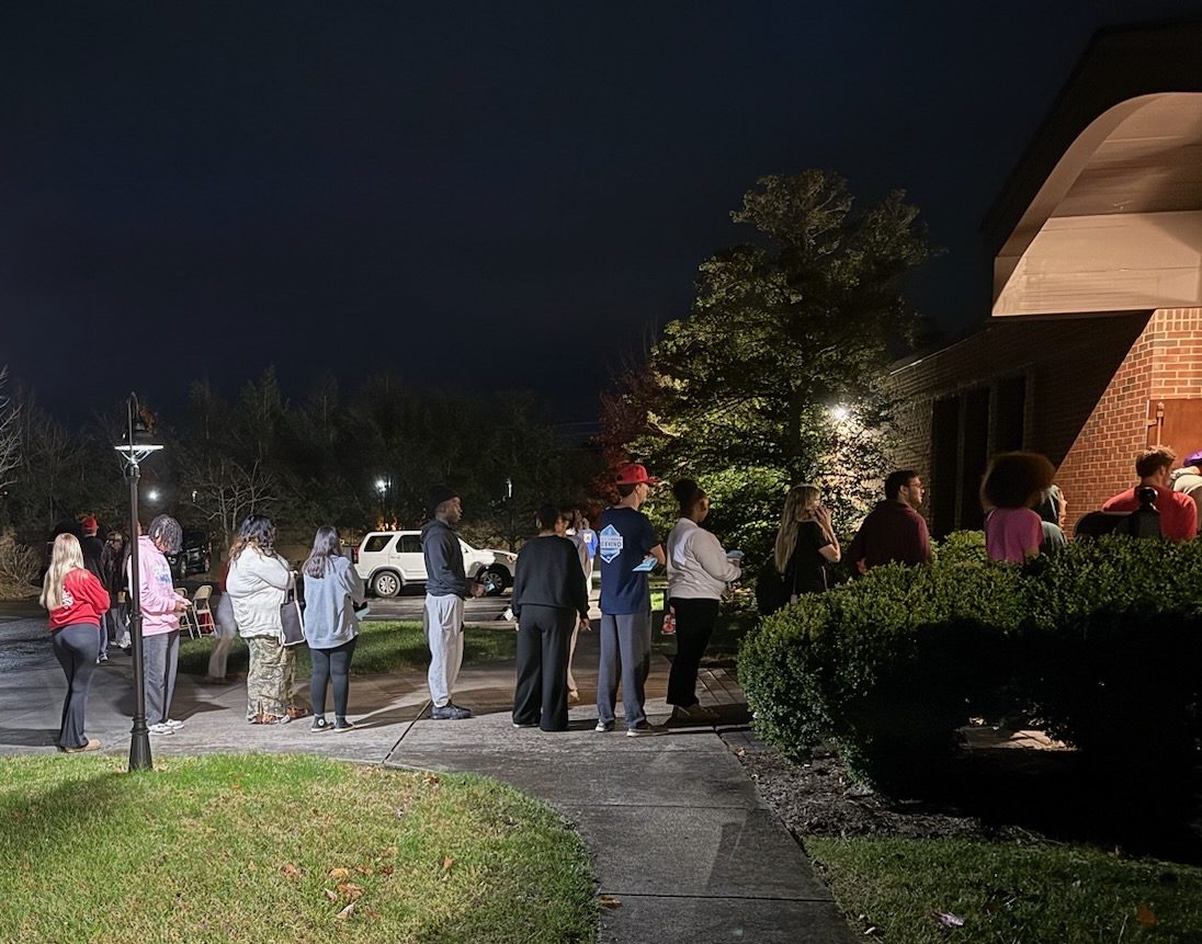 A line of people stretches from a building on the right side of the photo to the parking lot on the left side. The sky is dark.