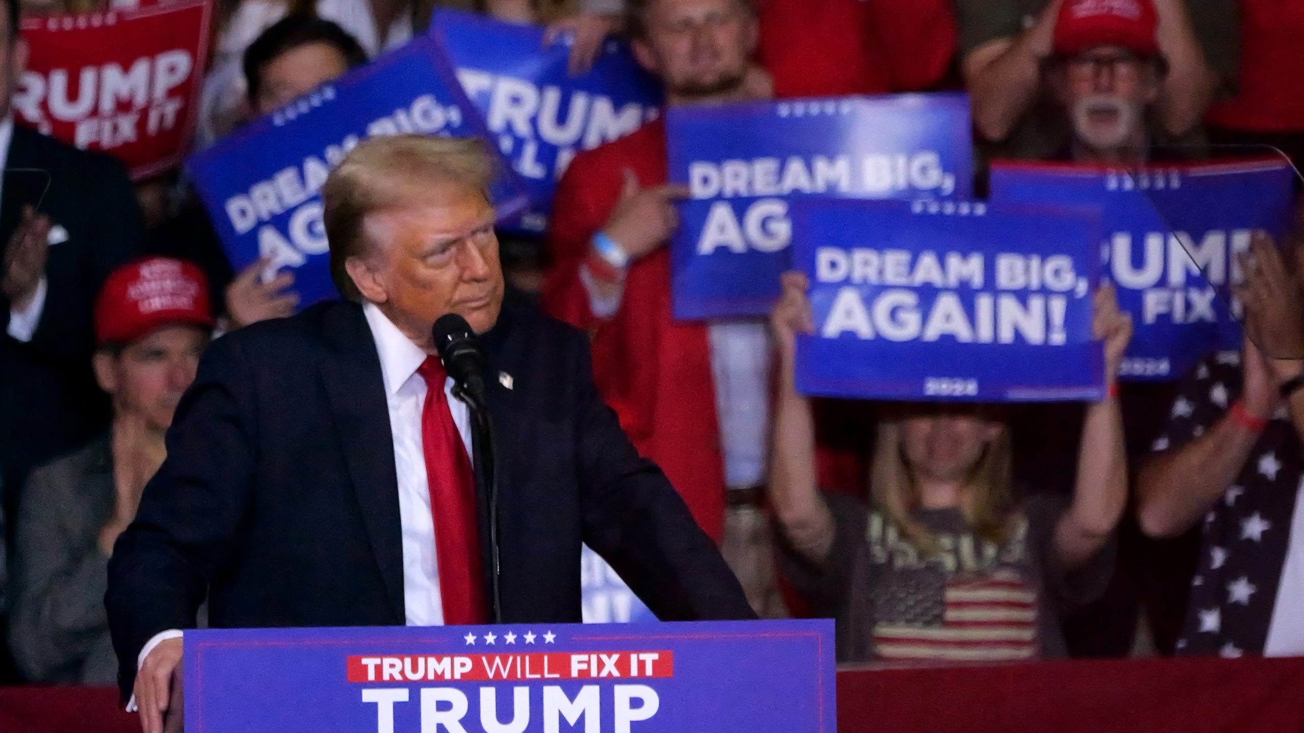 Former President Donald Trump stands at a podium surrounded by supporters with signs reading "Dream Big, Again!"