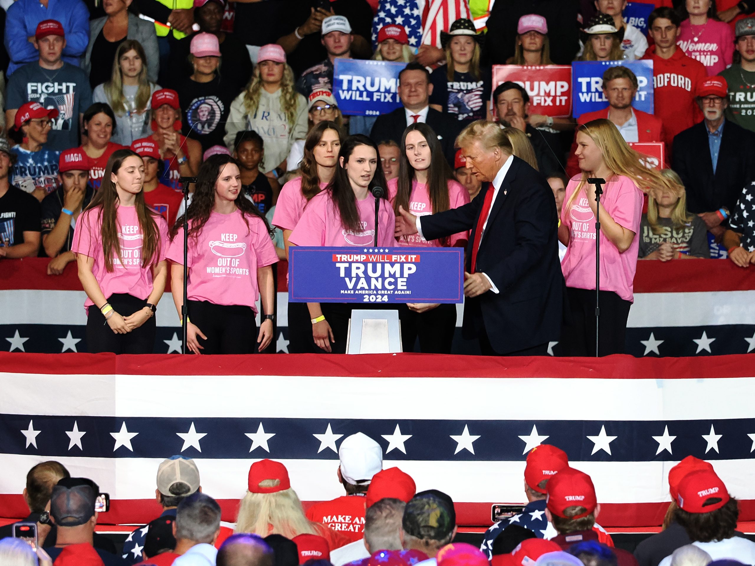 Women wearing t-shirts against transgender athletes stand with the former president at a political rally.