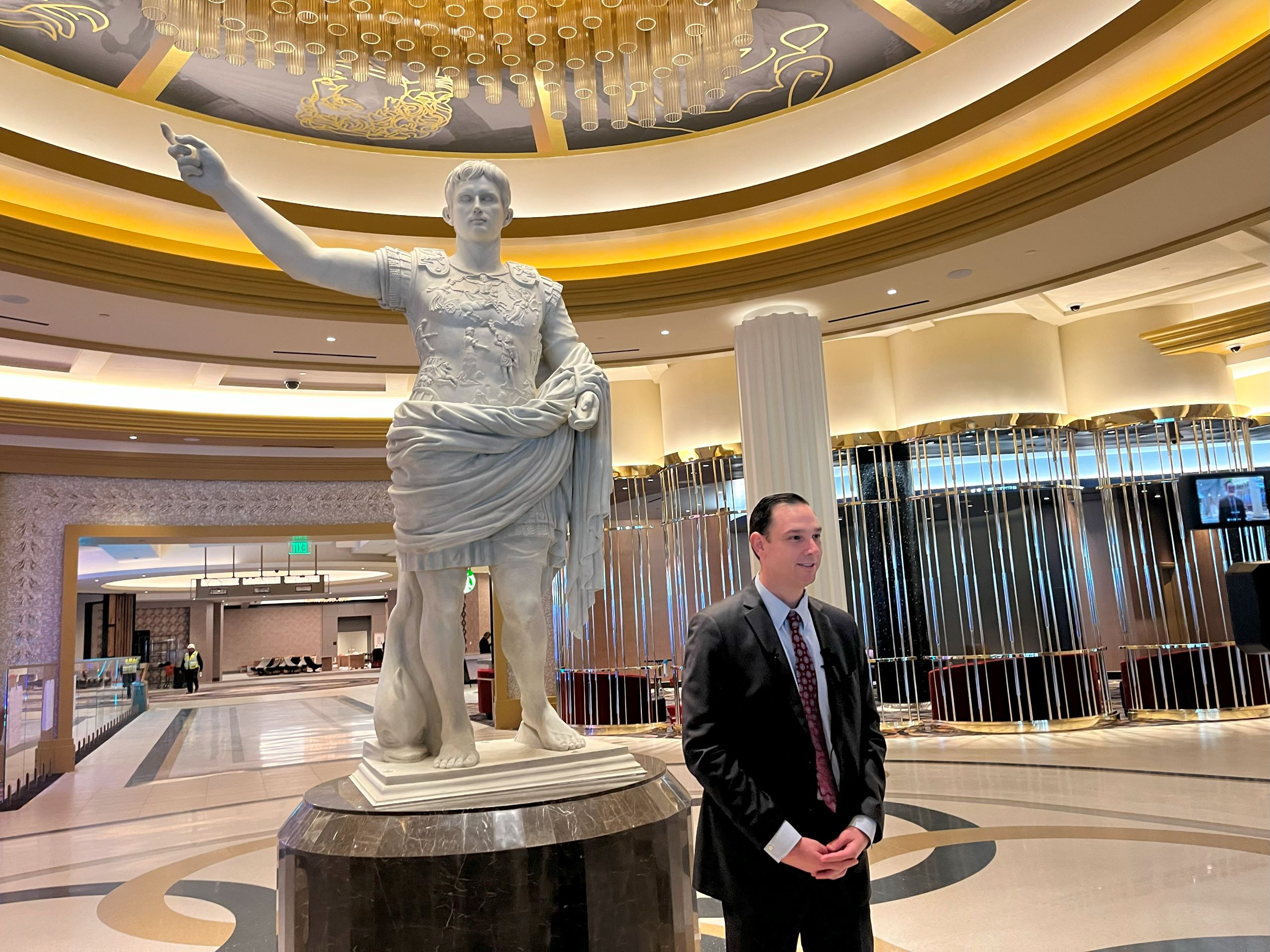 a man, Chris Albrecht with Caesars Virginia, stands in the casino resort lobby in front of a white marble statue of Augustus