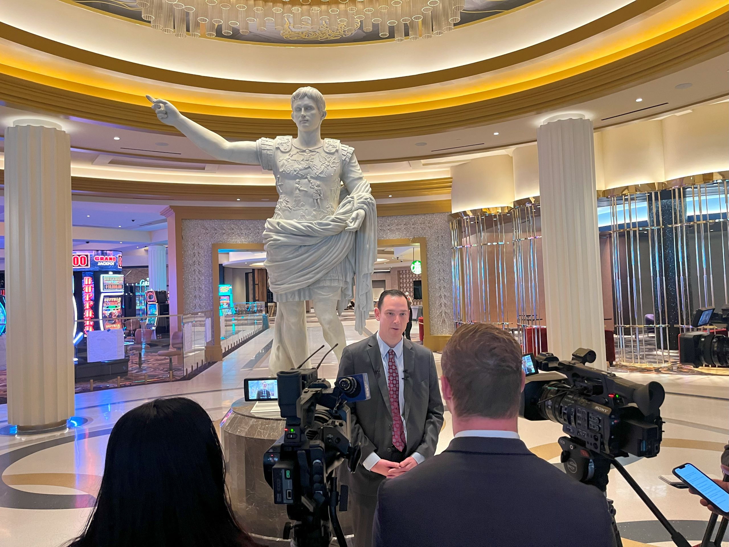 a man, Chris Albrecht with Caesars Virginia, stands in the casino resort lobby in front of a white marble statue of Augustus