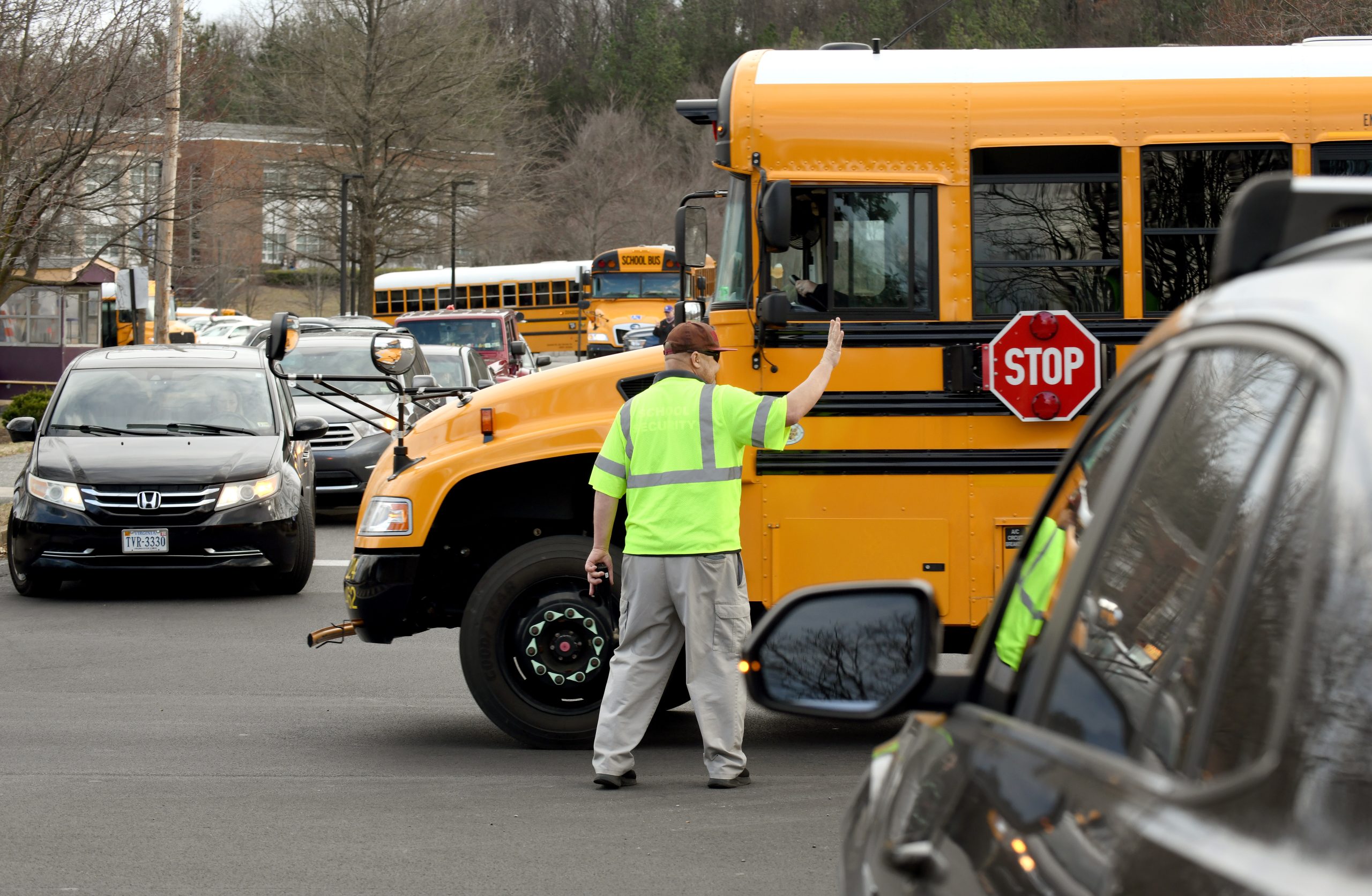 A man in a neon green vest directs traffic including a school bus and cars outside Patrick Henry High School