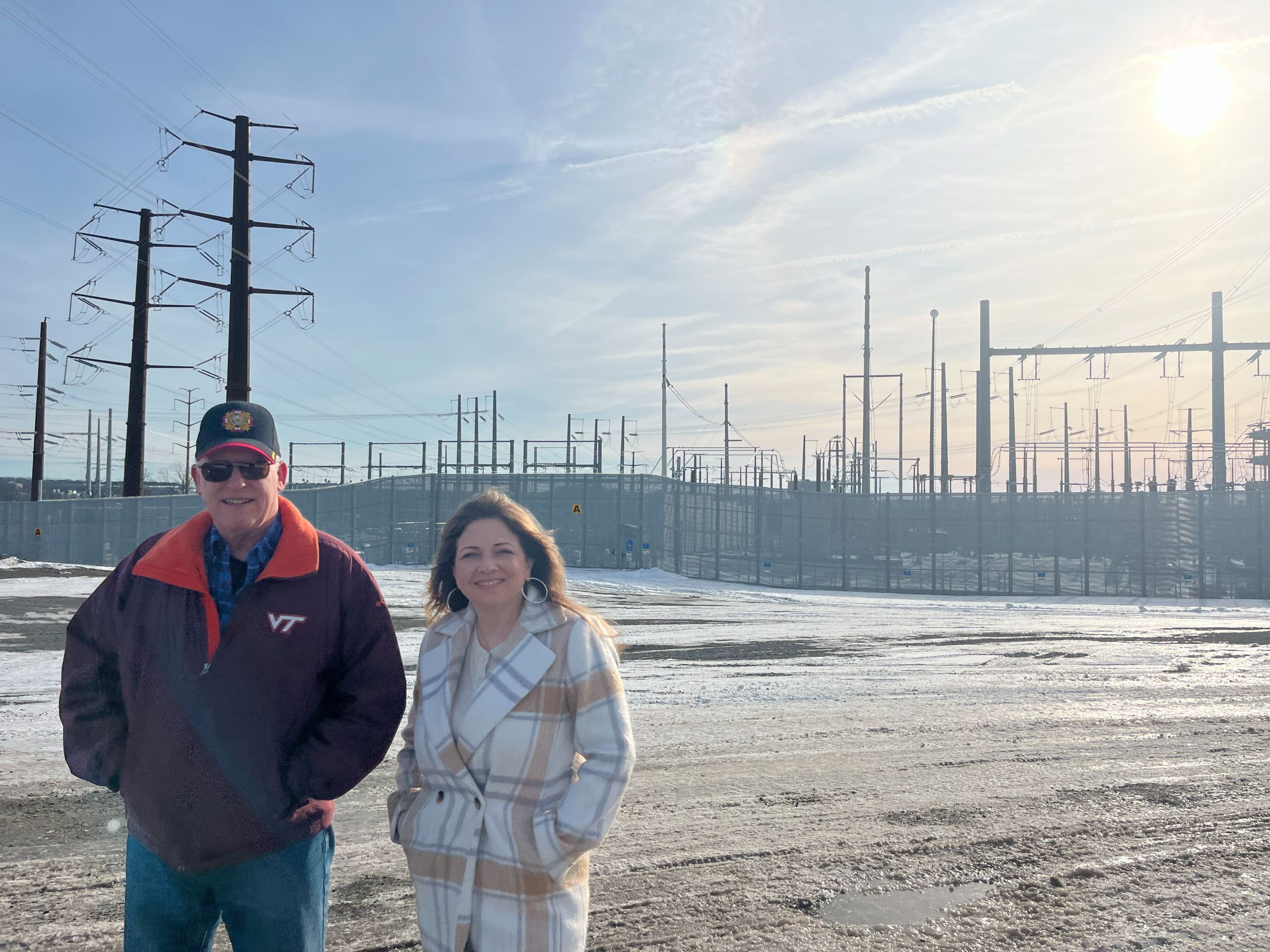 A man and a woman, Bill Wright and Elena Schlossberg, stand in front of a large substation in Loudoun County