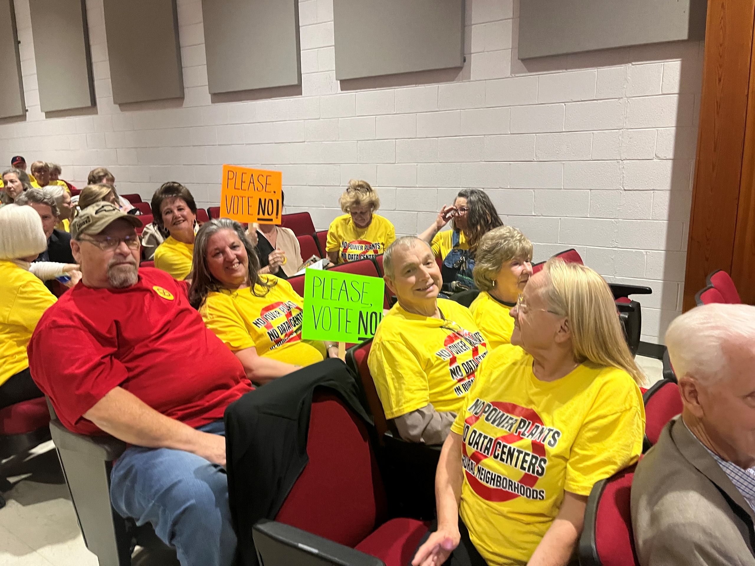 a group of Pittsylvania residents sit in auditorium seats, sporting t shirts and carrying signs to oppose a data center proposal