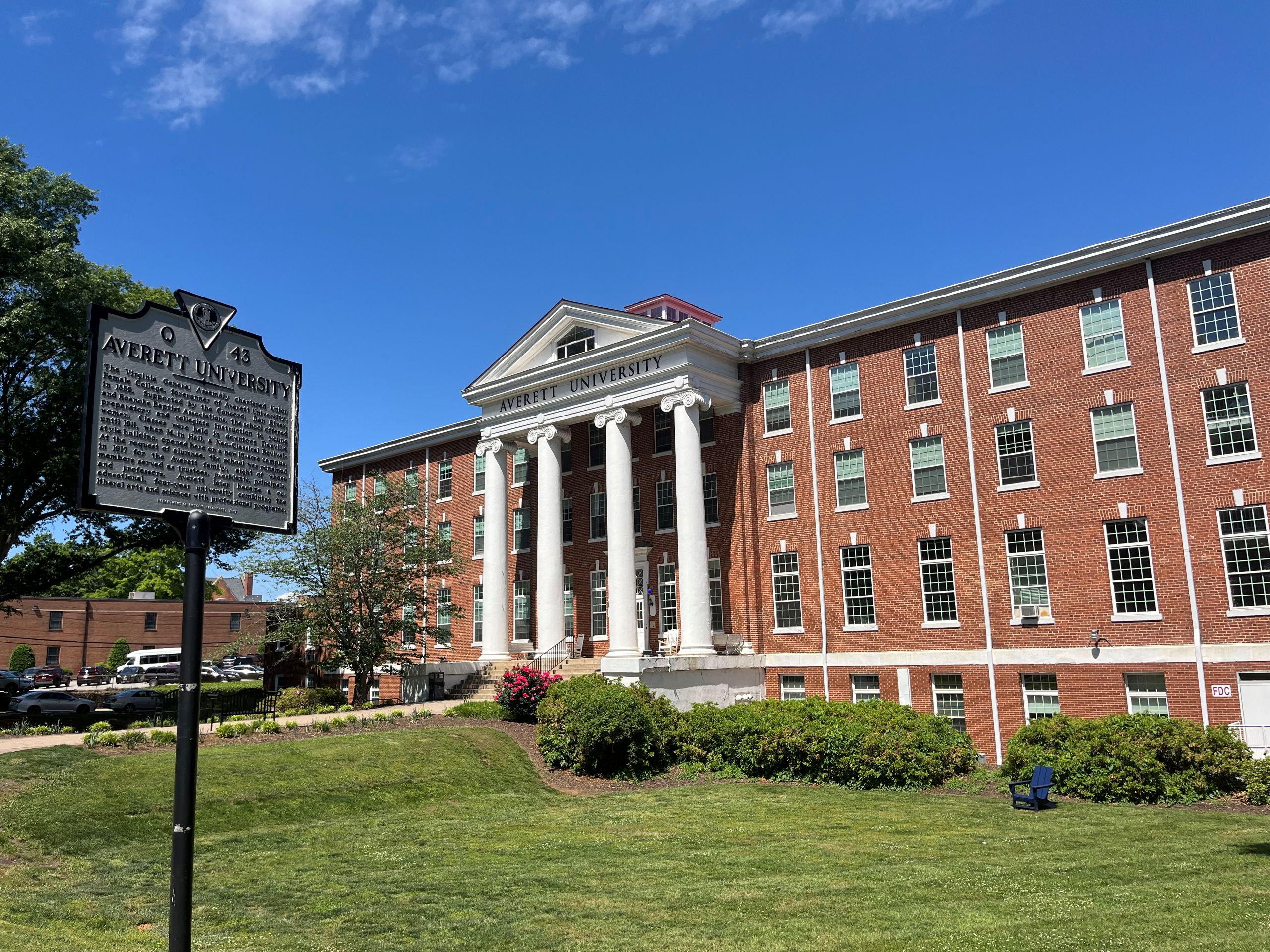a large brick building with many windows and white columns in front, the main hall at Averett University, stands behind a historic marker about the school